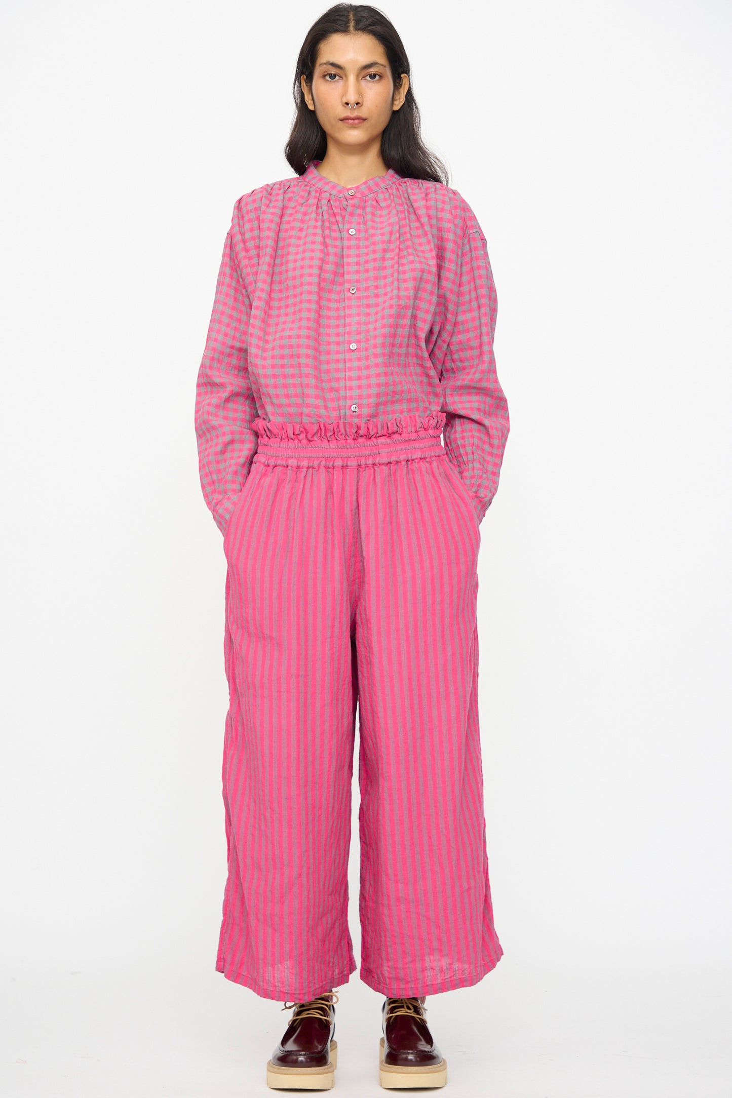 A person stands facing forward wearing Ichi Antiquités Linen Cotton Stripe Pants in Pink with an elastic waist, a loose-fitting pink and purple checkered long-sleeve top, and burgundy shoes.