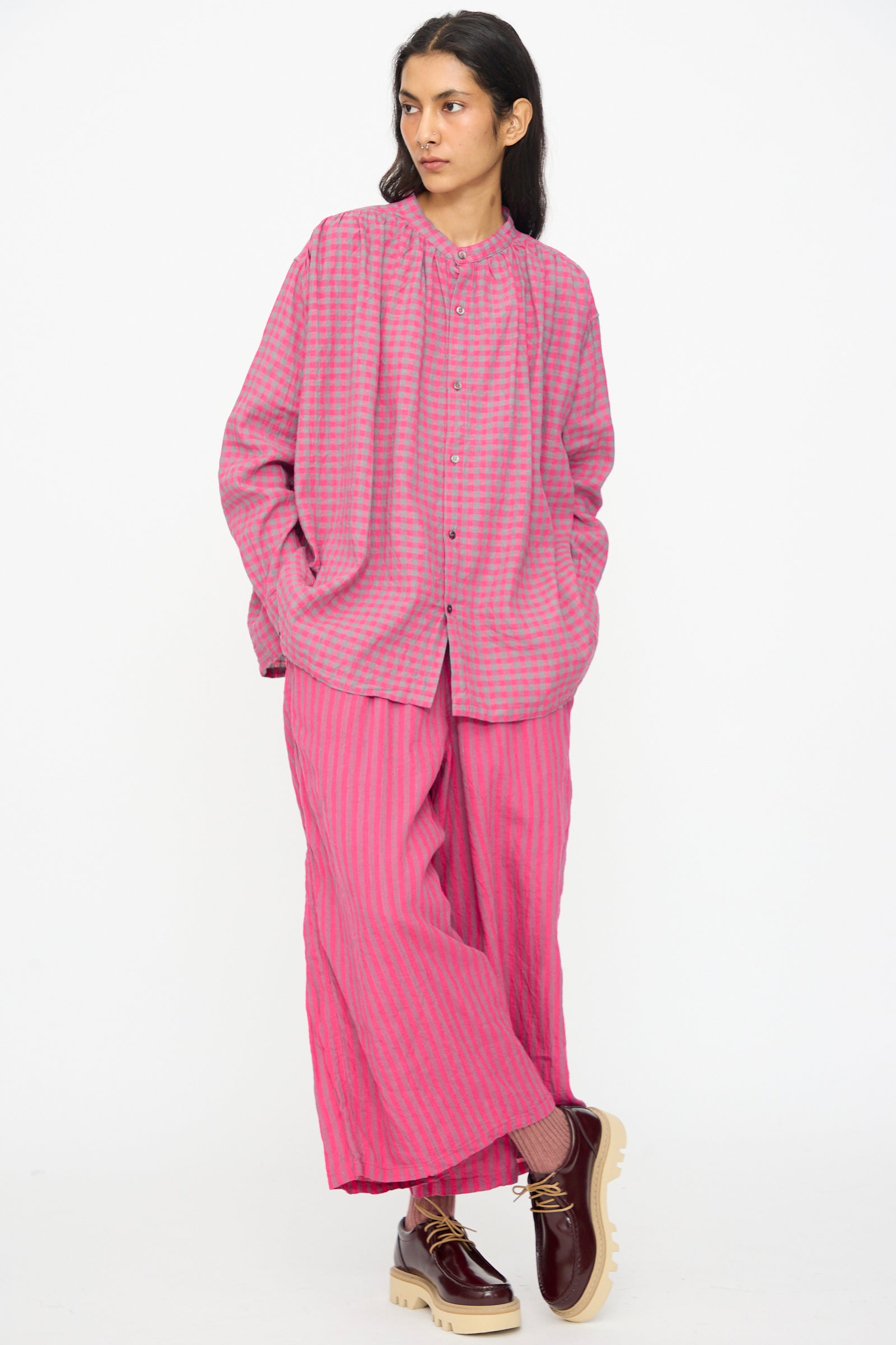 A person wears Ichi Antiquités Linen Cotton Stripe Pants in Pink with an elastic waist, a loose pink and gray checked shirt, and maroon platform shoes, standing against a plain white background.