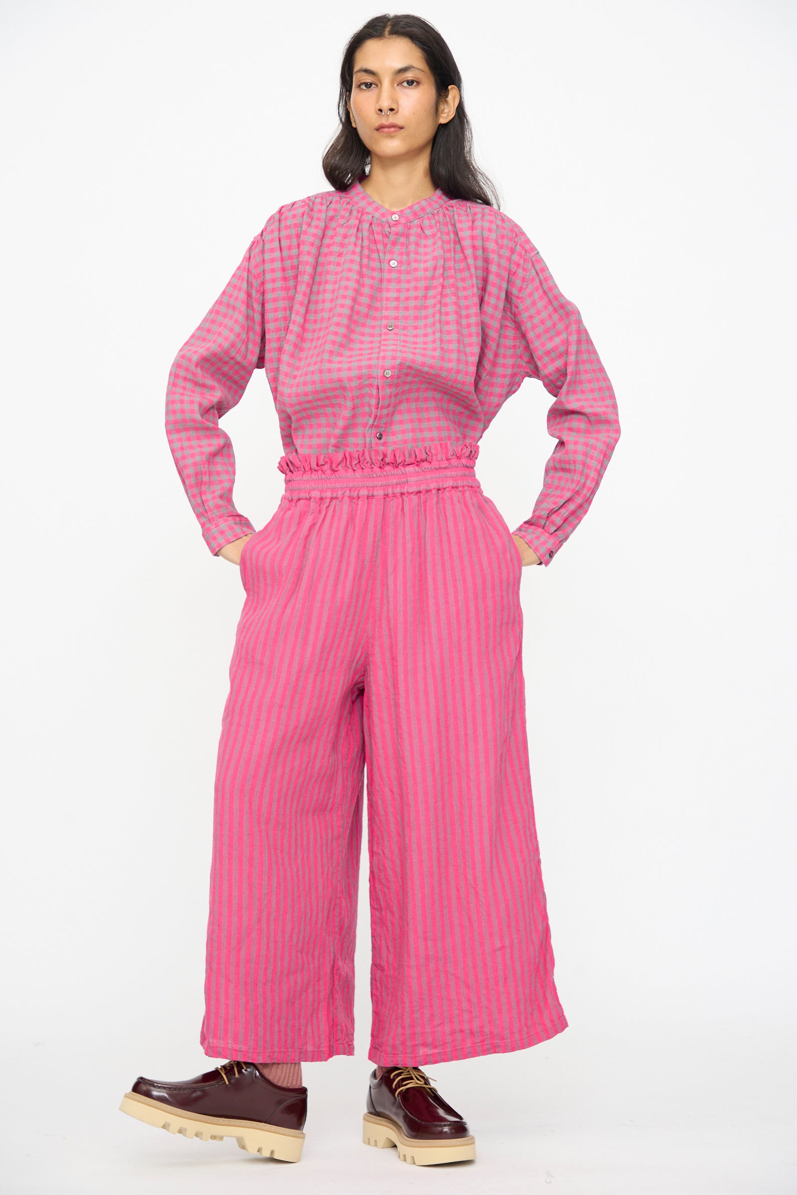 A person stands against a white background wearing Ichi Antiquités Linen Cotton Stripe Pants in Pink with an elasticated waist and cropped wide legs, paired with a matching long-sleeve shirt and maroon platform shoes.