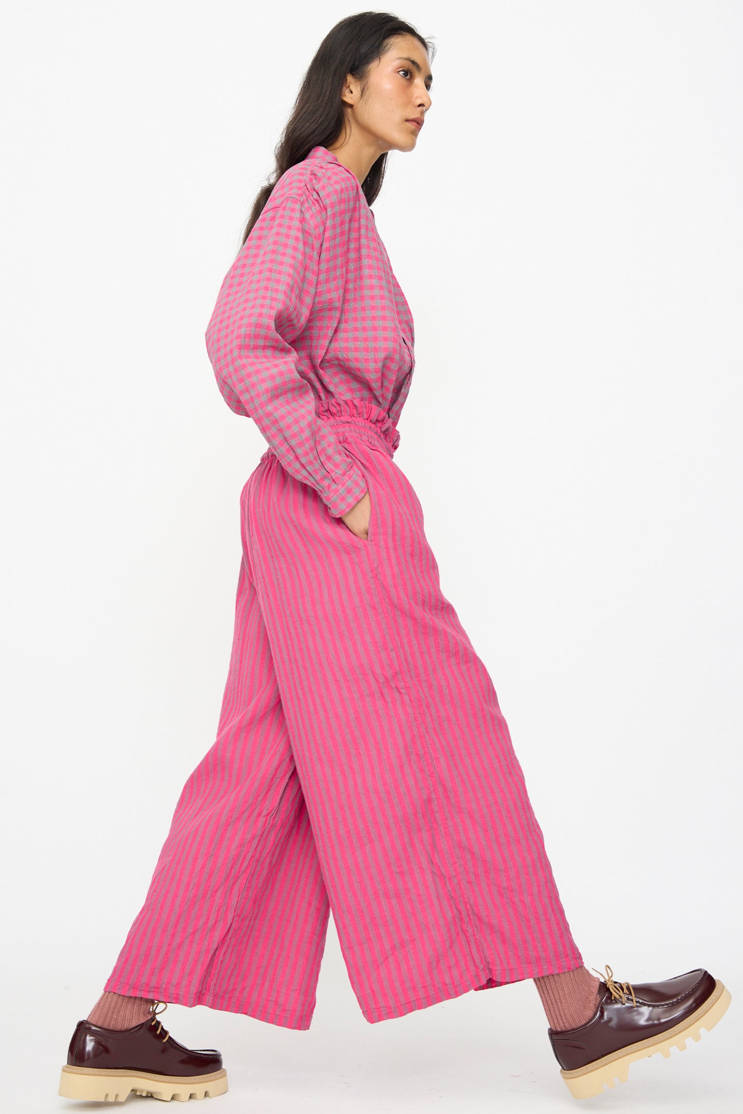 A woman wears Ichi Antiquités Linen Cotton Stripe Pants in Pink—loose, wide-leg, cropped with an elasticated waist—paired with brown platform shoes, walking against a plain white background.