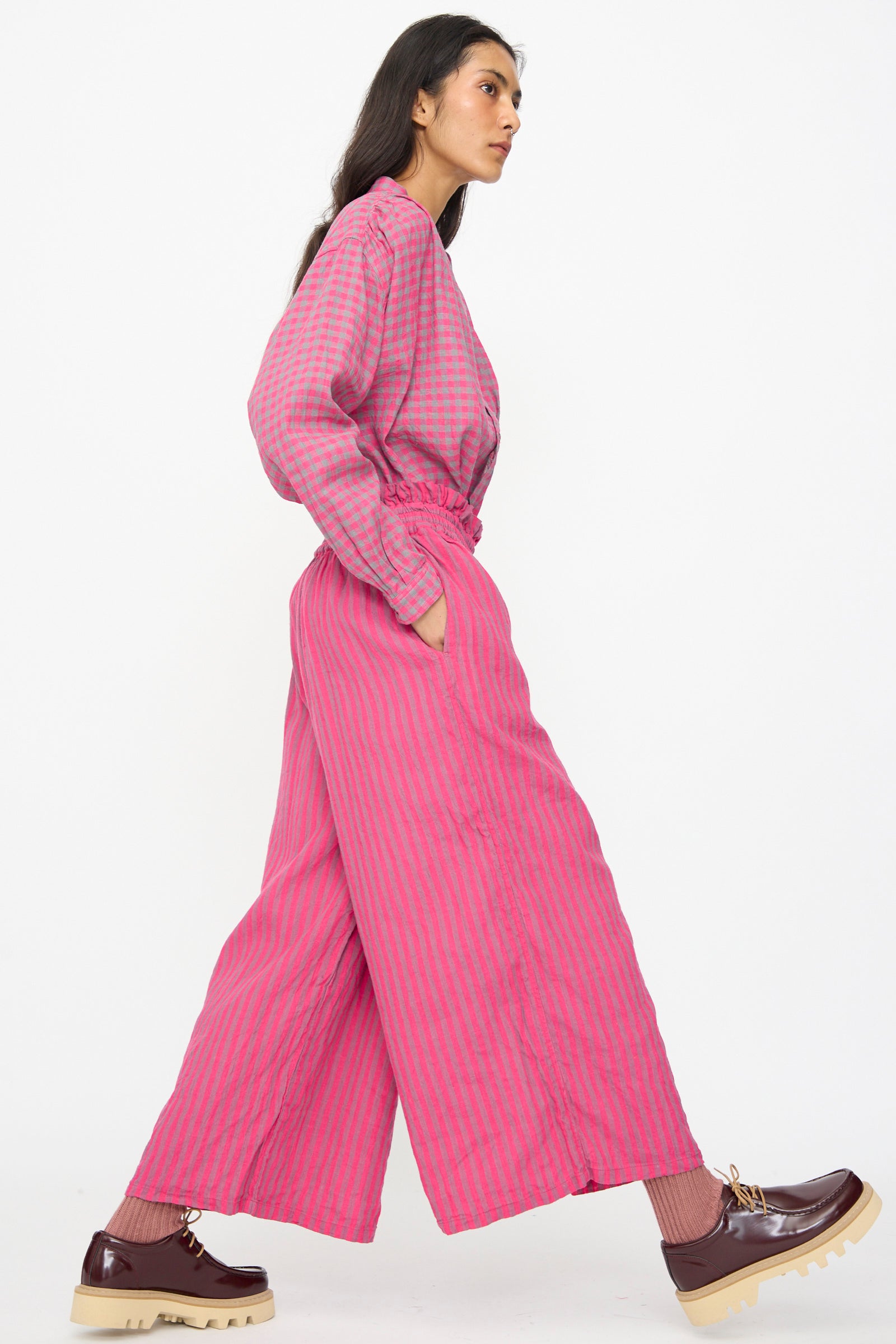 A woman wears Ichi Antiquités Linen Cotton Stripe Pants in Pink—loose, wide-leg, cropped with an elasticated waist—paired with brown platform shoes, walking against a plain white background.