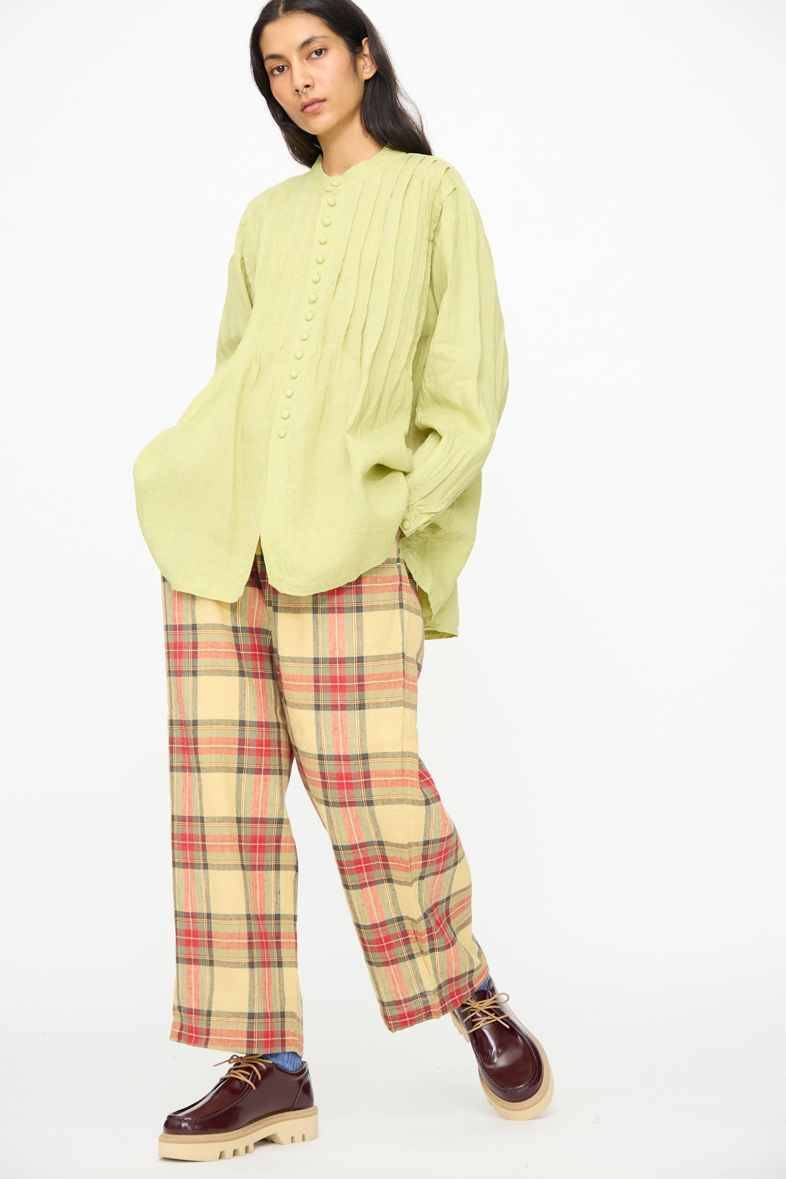 A person stands against a plain white background wearing Ichi Antiquités Linen Cotton Tartan Pants in Butter Yellow, paired with a light green buttoned blouse and burgundy platform shoes.