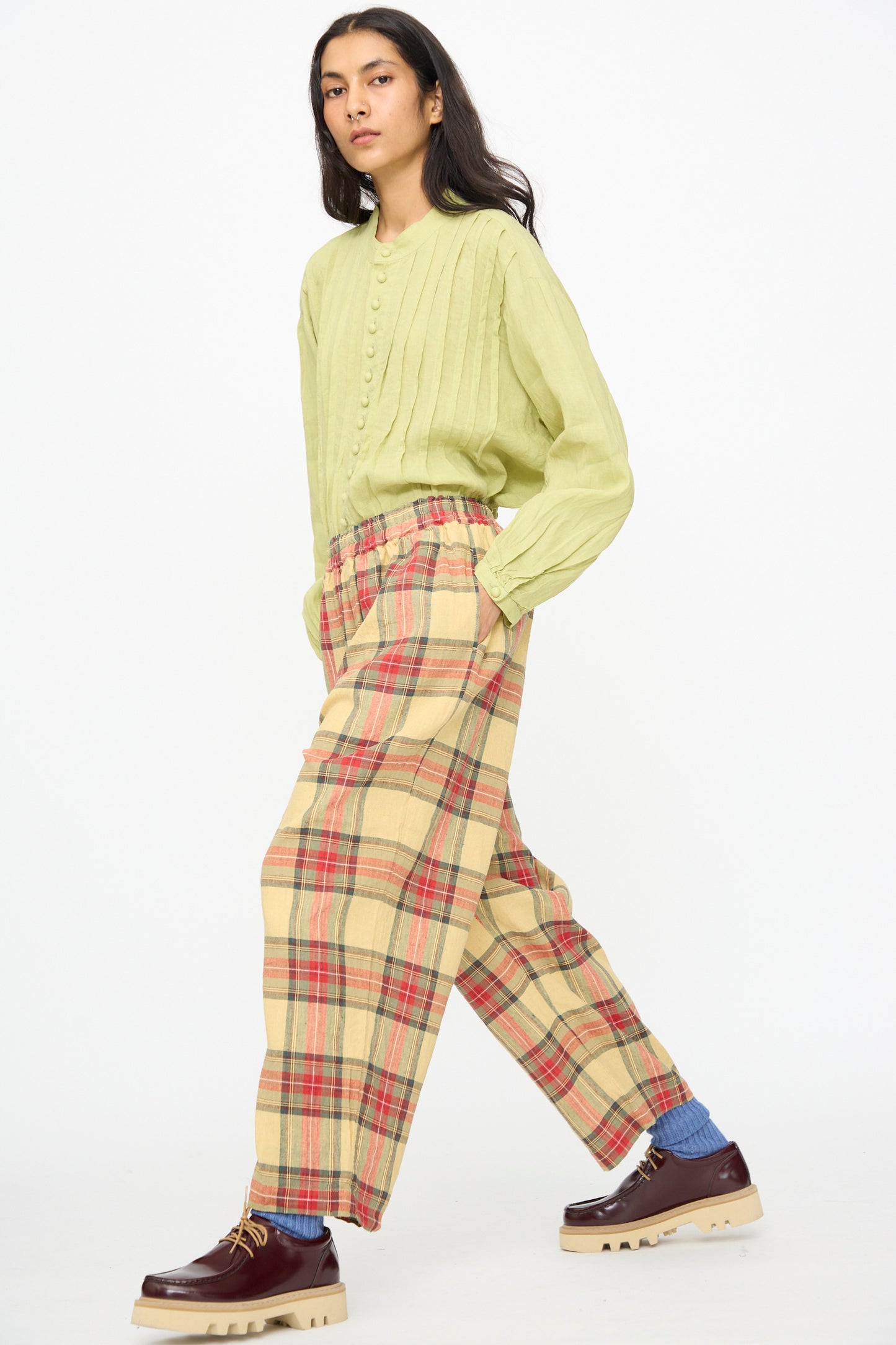 A person wearing a light green buttoned shirt, Ichi Antiquités Linen Cotton Tartan Pants in Butter Yellow, brown shoes, and blue socks stands against a plain white background.