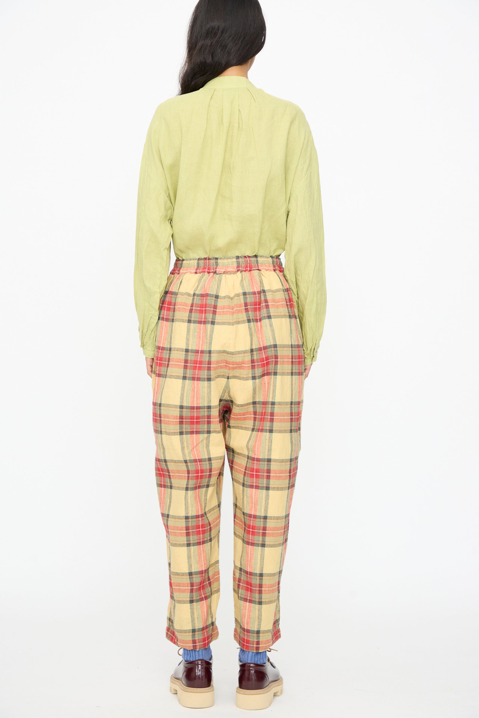 A person stands facing away, wearing a light green long-sleeve shirt and Ichi Antiquités Linen Cotton Tartan Pants in Butter Yellow—high-waisted, wide leg, made in Japan—with red and yellow stripes, brown shoes, and visible blue socks.