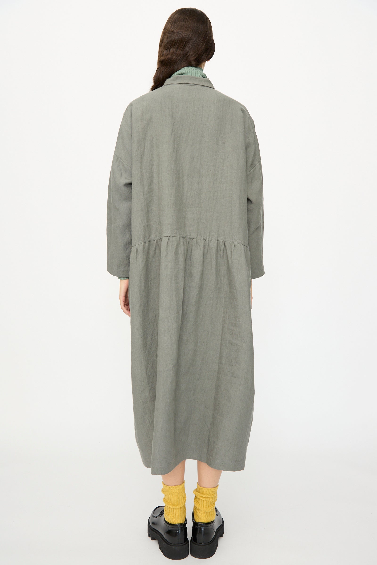 A person stands facing away, wearing the Ichi Antiquités Linen Dress in Khaki with yellow socks and black platform shoes against a plain white background. This elegant long sleeve, gathered waist dress is made in Japan.