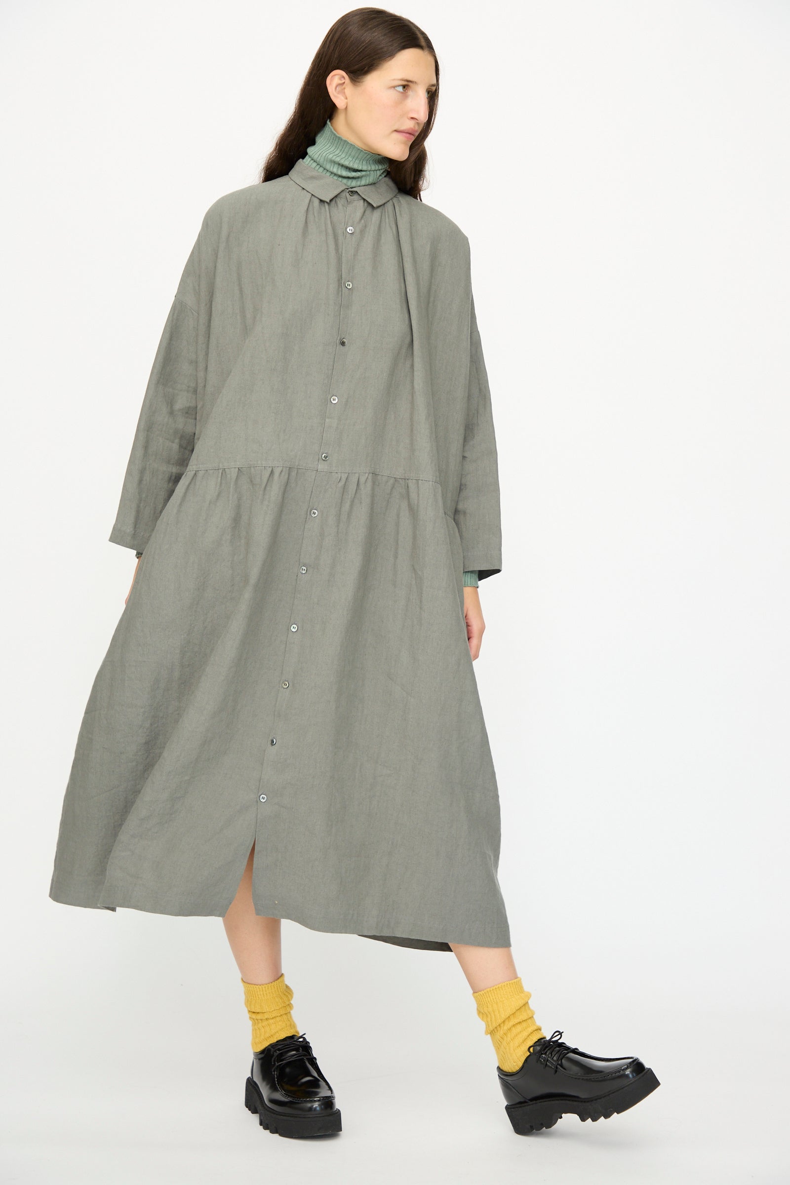 A woman stands against a plain white background wearing the Ichi Antiquités Linen Dress in Khaki, styled over a green turtleneck, with yellow socks and black platform shoes.