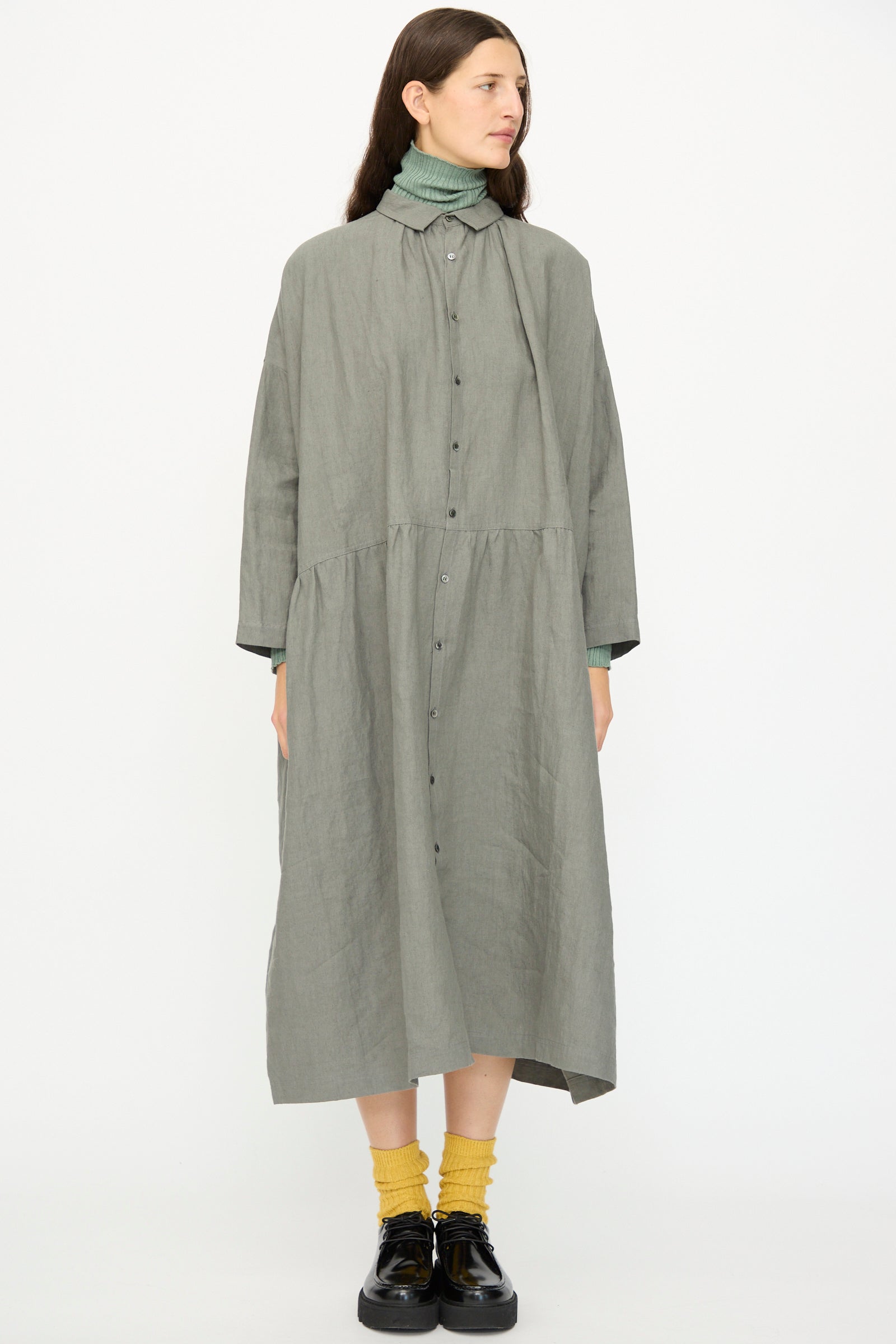 A woman wears the Ichi Antiquités Linen Dress in Khaki over a green turtleneck, paired with yellow socks and black shoes, standing against a plain white background.