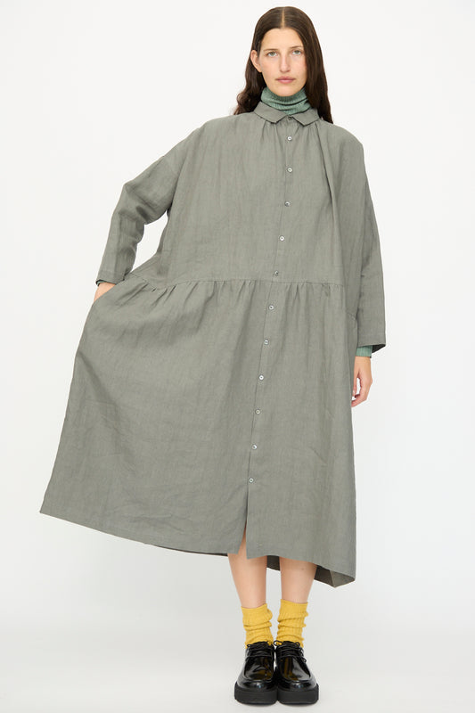 A person wears the Ichi Antiquités Linen Dress in Khaki—loose-fitting, long sleeve, made in Japan—over a green turtleneck, paired with yellow socks and black shoes, standing against a plain white background.