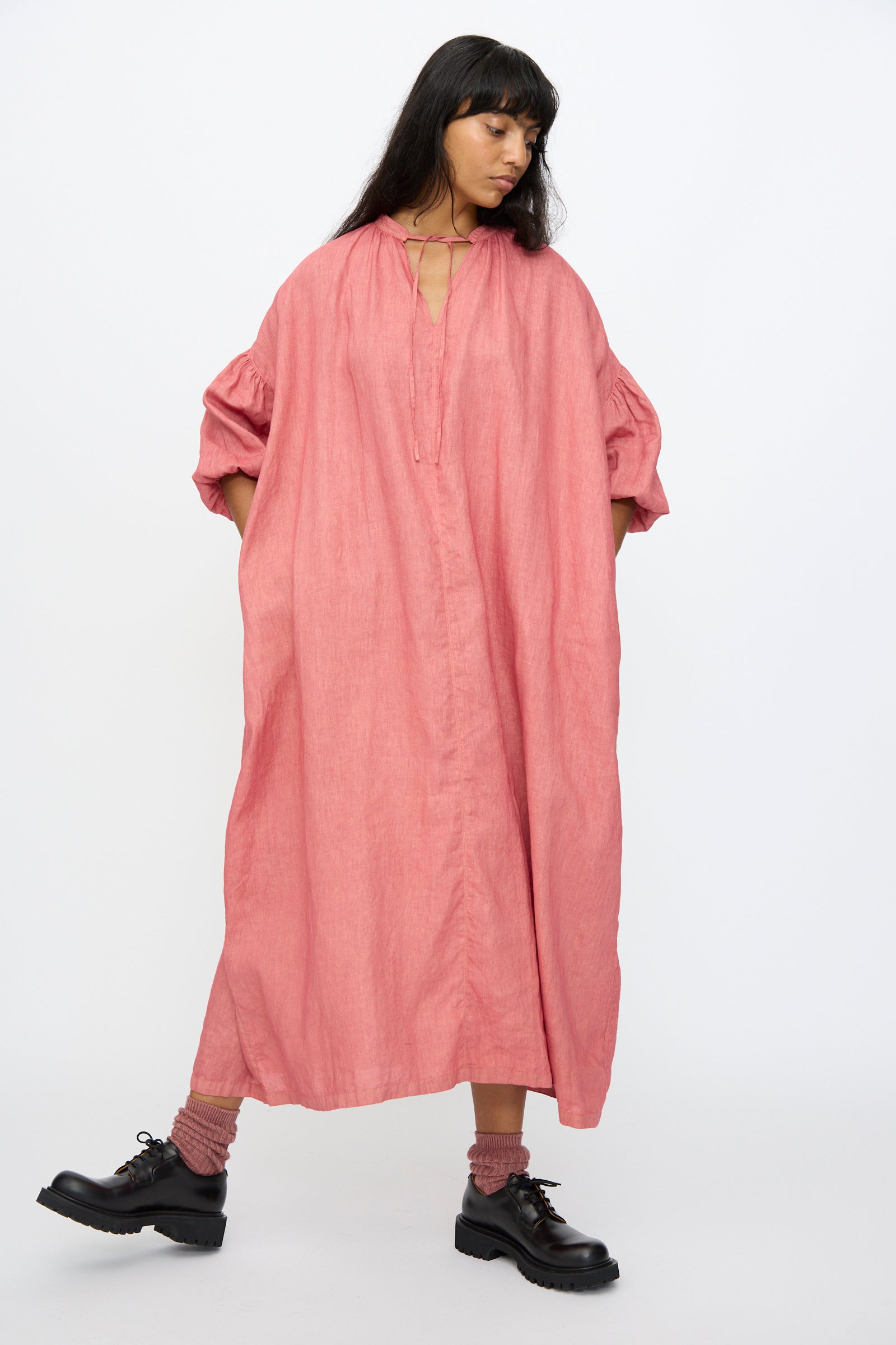 A woman stands against a white background wearing the Ichi Antiquités Linen Dress in Pink—an oversized, hand-dyed maxi with puffed sleeves—paired with black shoes and matching pink socks.