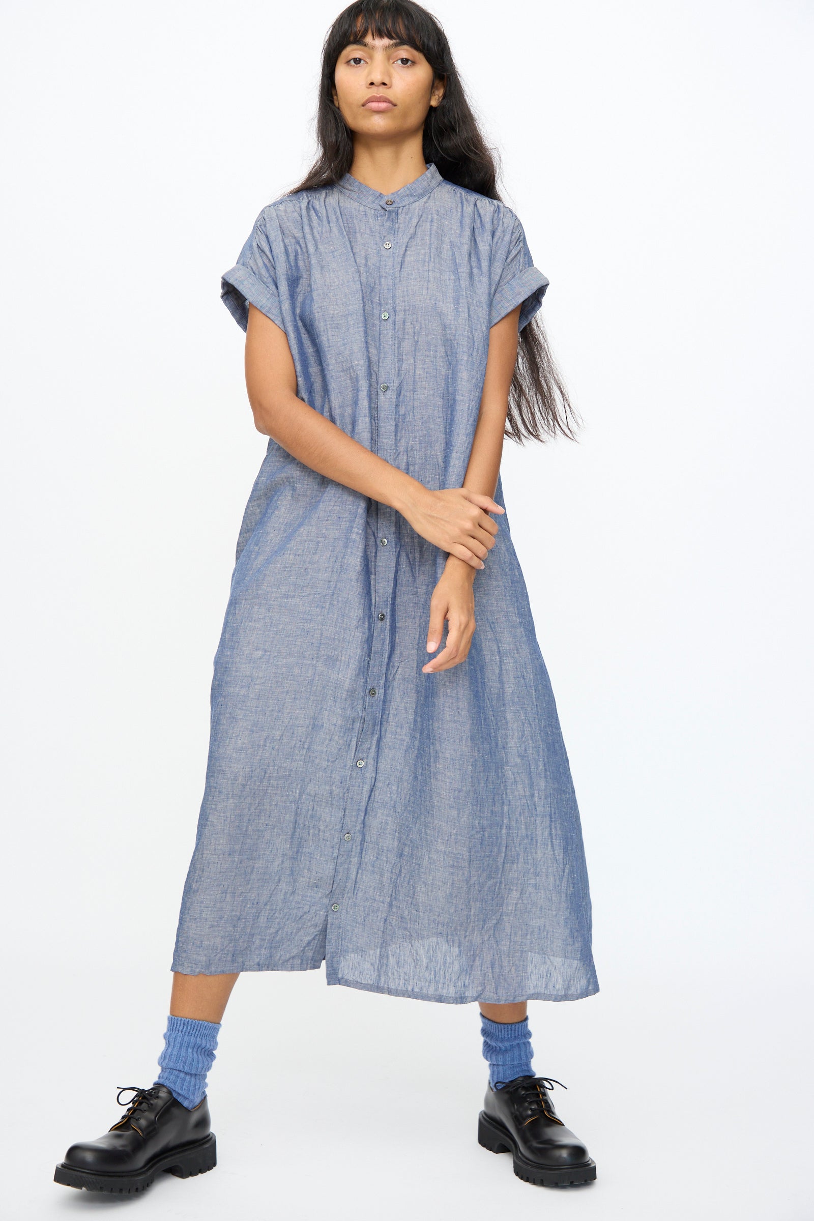 A person stands against a white background, wearing the Ichi Antiquités Linen Dungaree Dress in Blue—loose fit, short sleeves, button-down—paired with blue socks and black shoes.