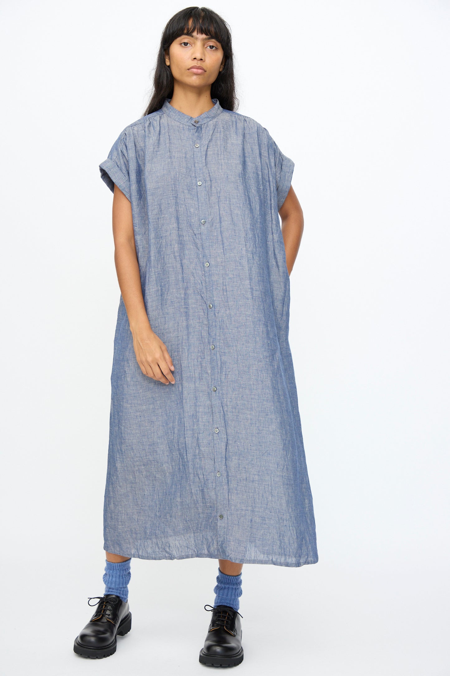 A person stands against a plain background wearing the Ichi Antiquités Linen Dungaree Dress in Blue, featuring a relaxed fit and ankle length, paired with blue socks and black shoes.
