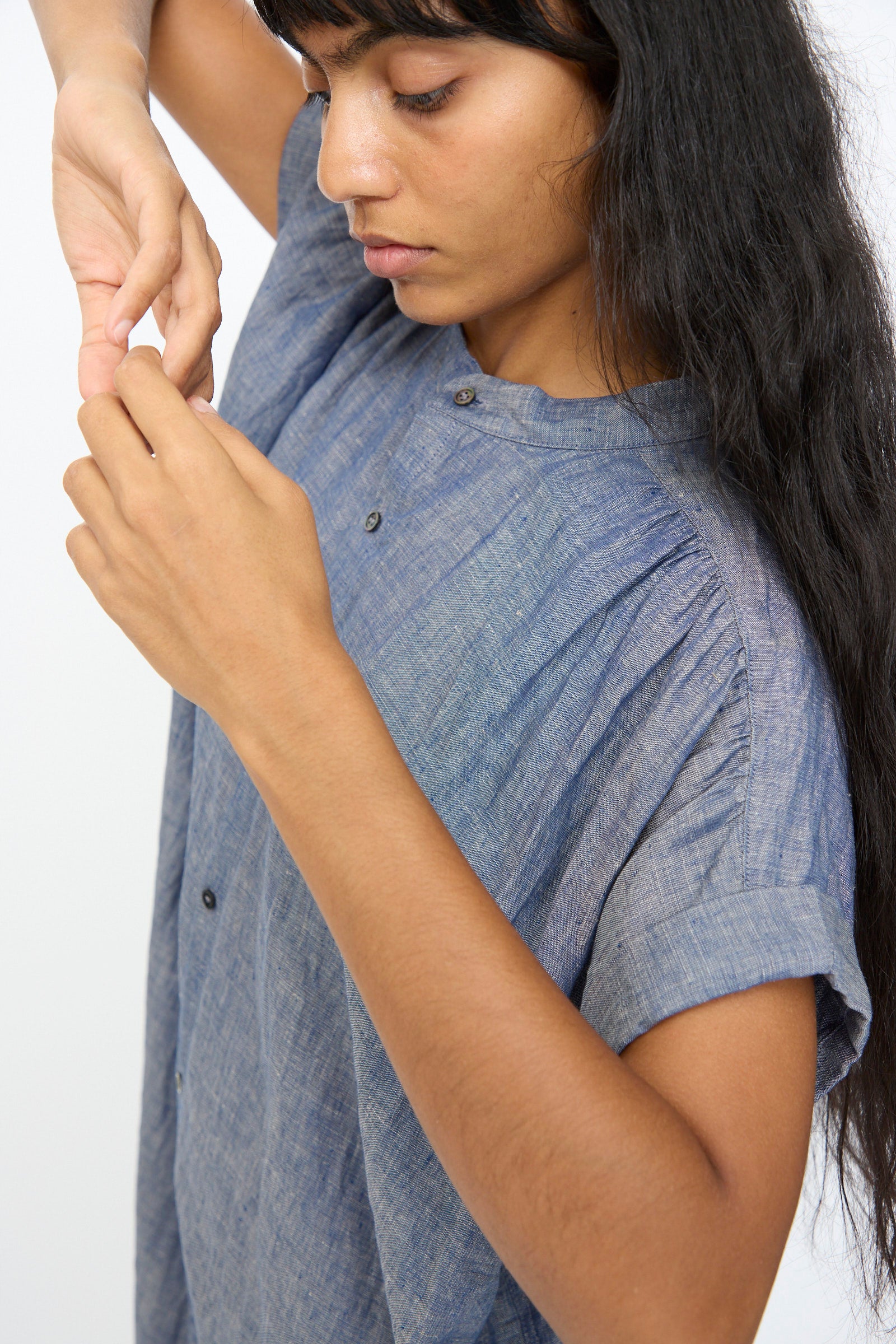 Wearing the Ichi Antiquités Linen Dungaree Dress in Blue, a person with long dark hair adjusts their fingers and looks down, set against a plain light background.