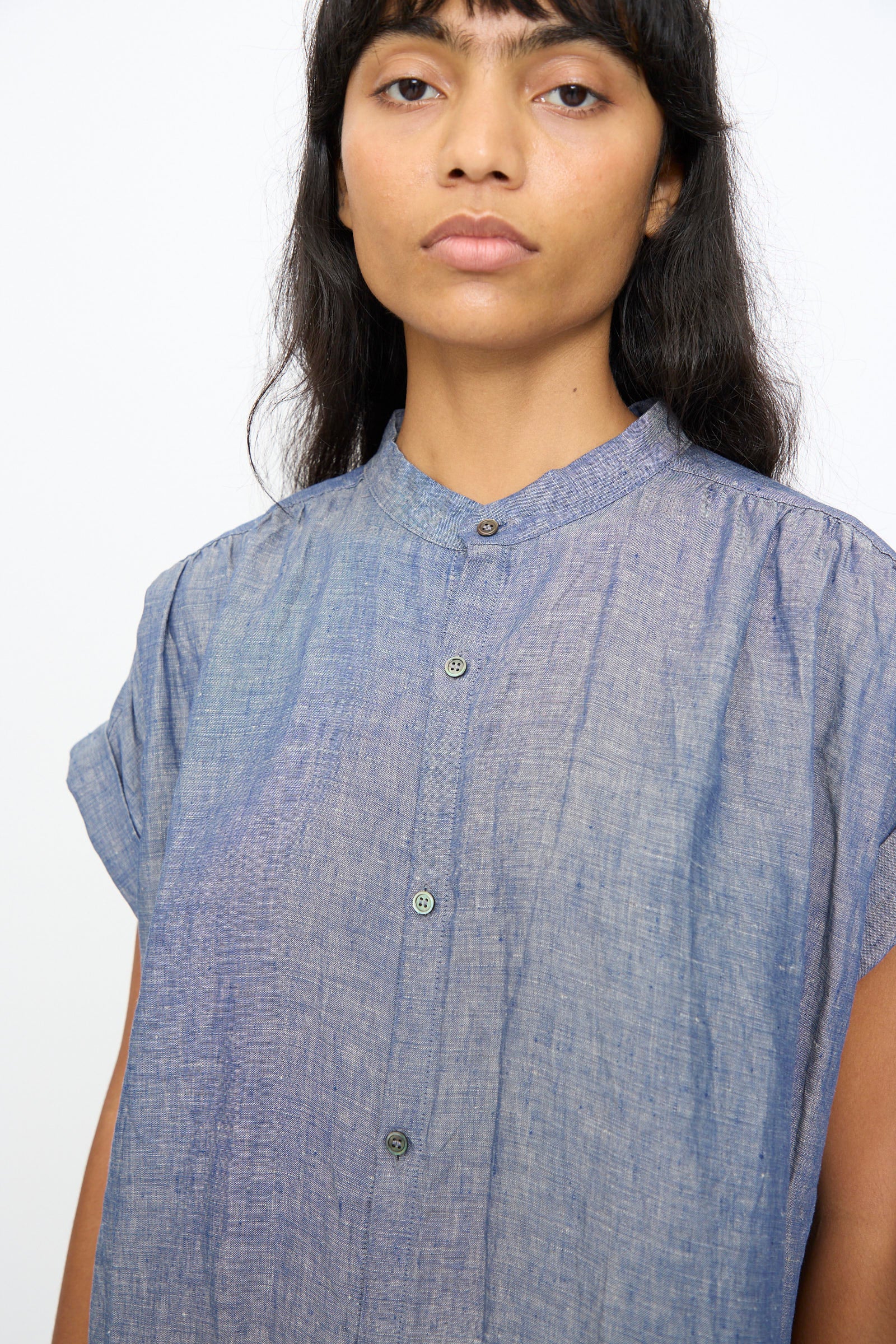 A person with long dark hair wears the Ichi Antiquités Linen Dungaree Dress in Blue, featuring short sleeves, a loose silhouette, and a mandarin collar, standing against a plain light background.