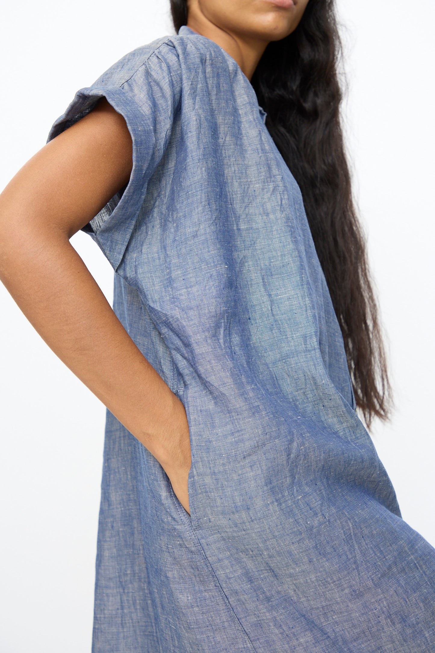 A person with long dark hair wears the Ichi Antiquités Linen Dungaree Dress in Blue, a relaxed fit, short-sleeved style, with a hand in the pocket, standing against a plain background.