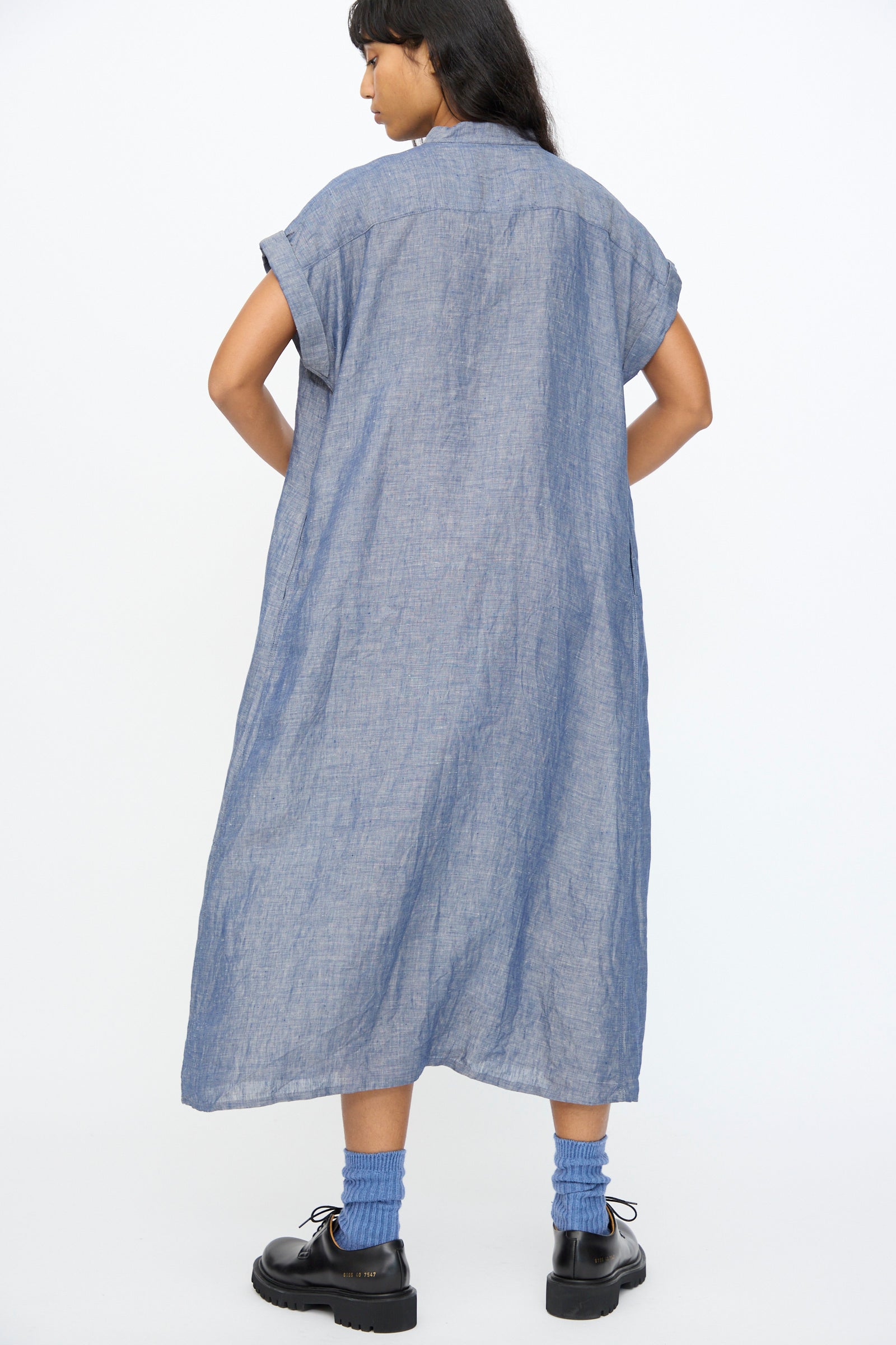 A woman faces away, wearing the Ichi Antiquités Linen Dungaree Dress in Blue with short sleeves, blue socks, and black shoes against a plain white background.