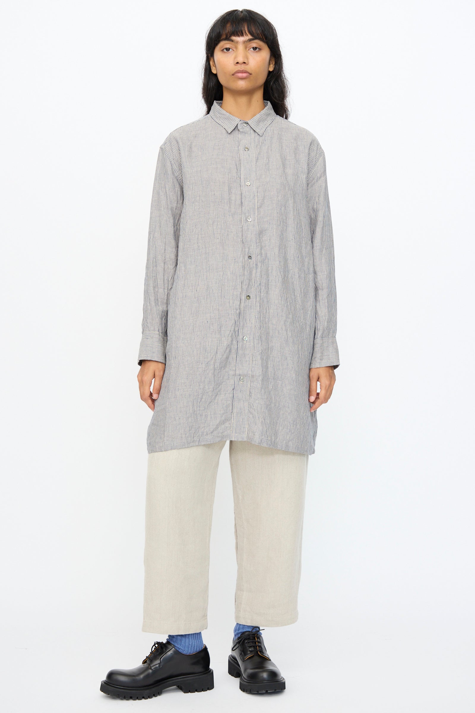 A person stands facing forward in the Ichi Antiquités Linen Dungaree Shirt in Stripe, styled with light beige cropped pants, blue socks, and black shoes against a plain white background.