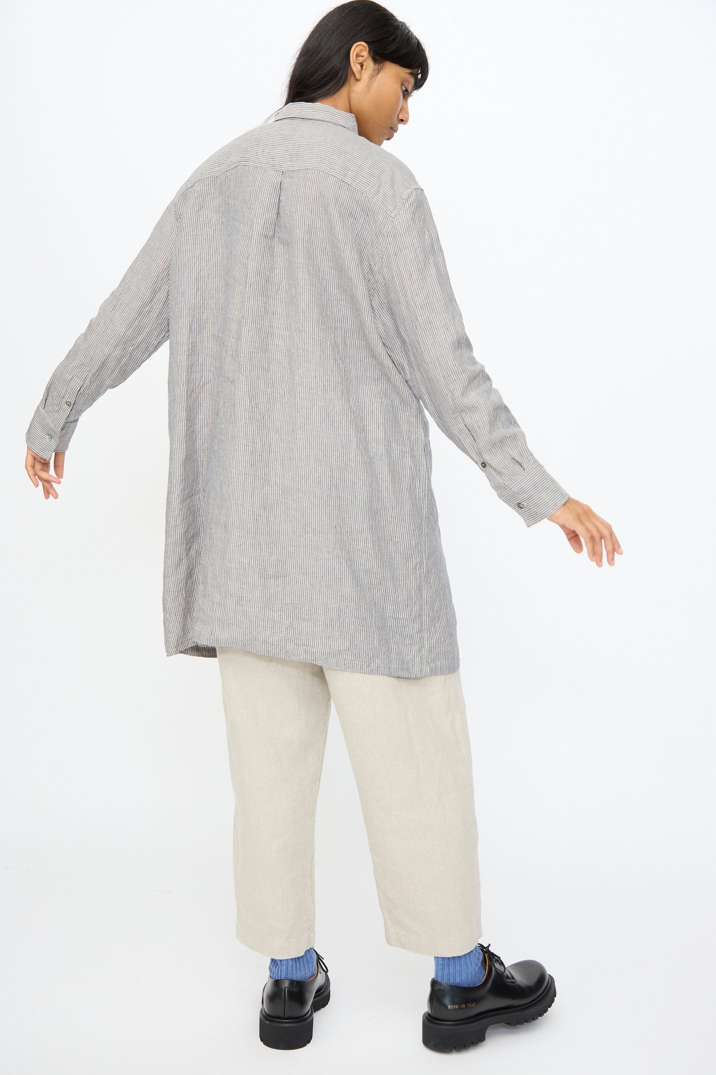 Against a plain white background, a person faces away wearing the Ichi Antiquités Linen Dungaree Shirt in Stripe, paired with light beige cropped pants, blue socks, and black shoes.