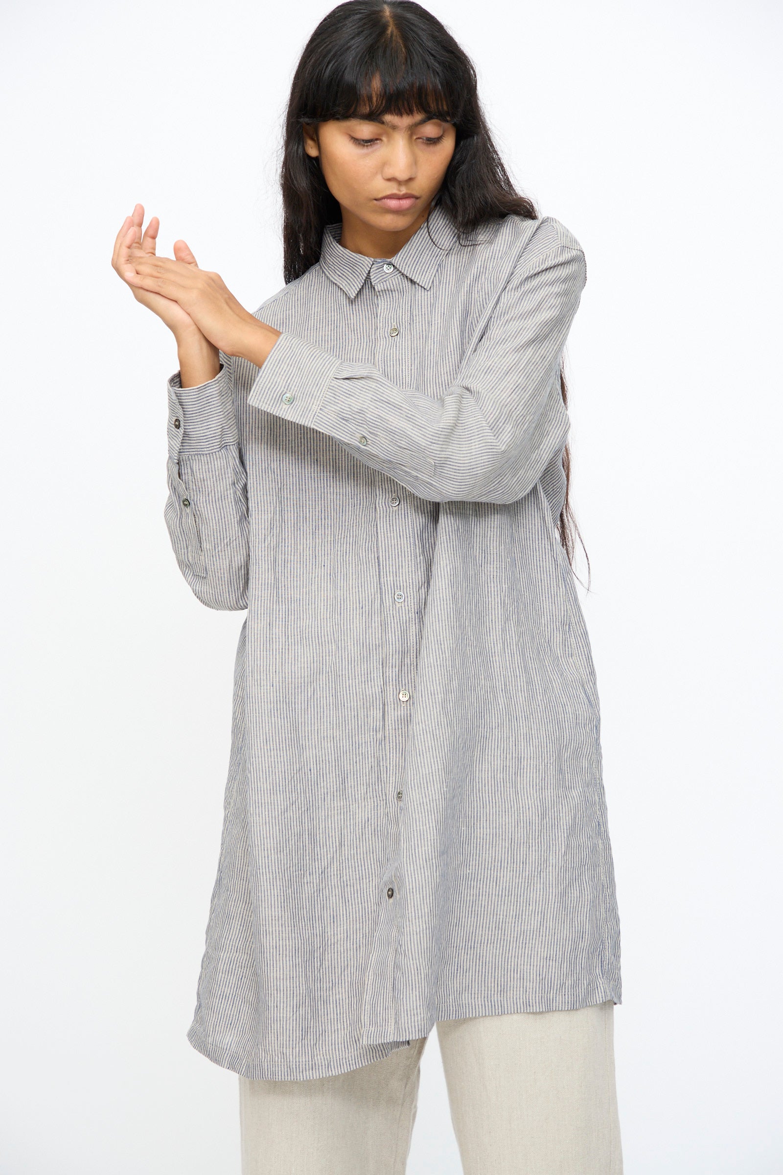 A woman in the Ichi Antiquités Linen Dungaree Shirt in Stripe— a long, gray, 100% linen shirt with thin vertical stripes—stands against a plain white background, paired with light-colored pants.