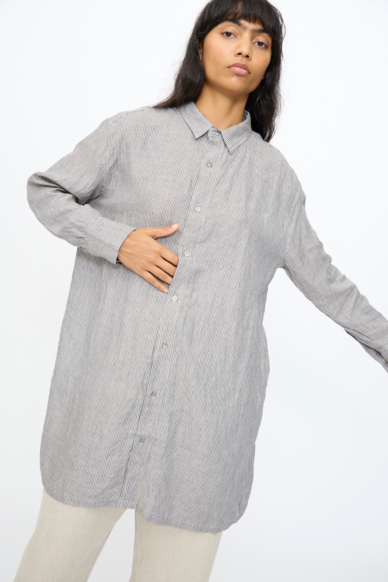 A person wears the Ichi Antiquités Linen Dungaree Shirt in Stripe, a long, button-up 100% linen shirt in light gray, paired with beige pants, standing against a plain white background.