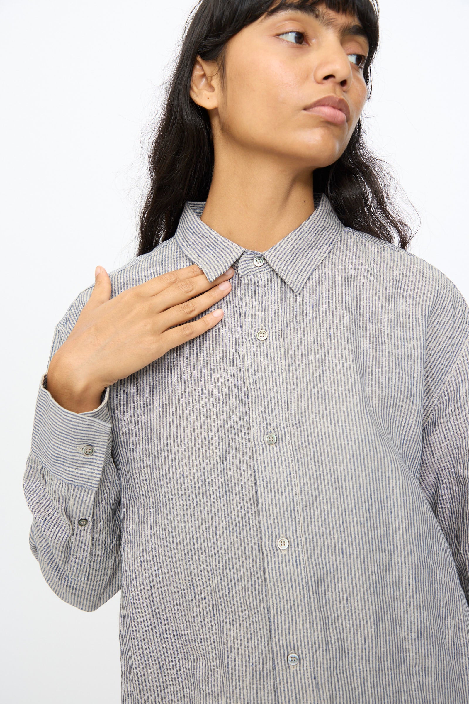 A person wearing the Ichi Antiquités Linen Dungaree Shirt in Stripe, with thin vertical stripes, touches their chest with one hand and looks to the side against a plain background.