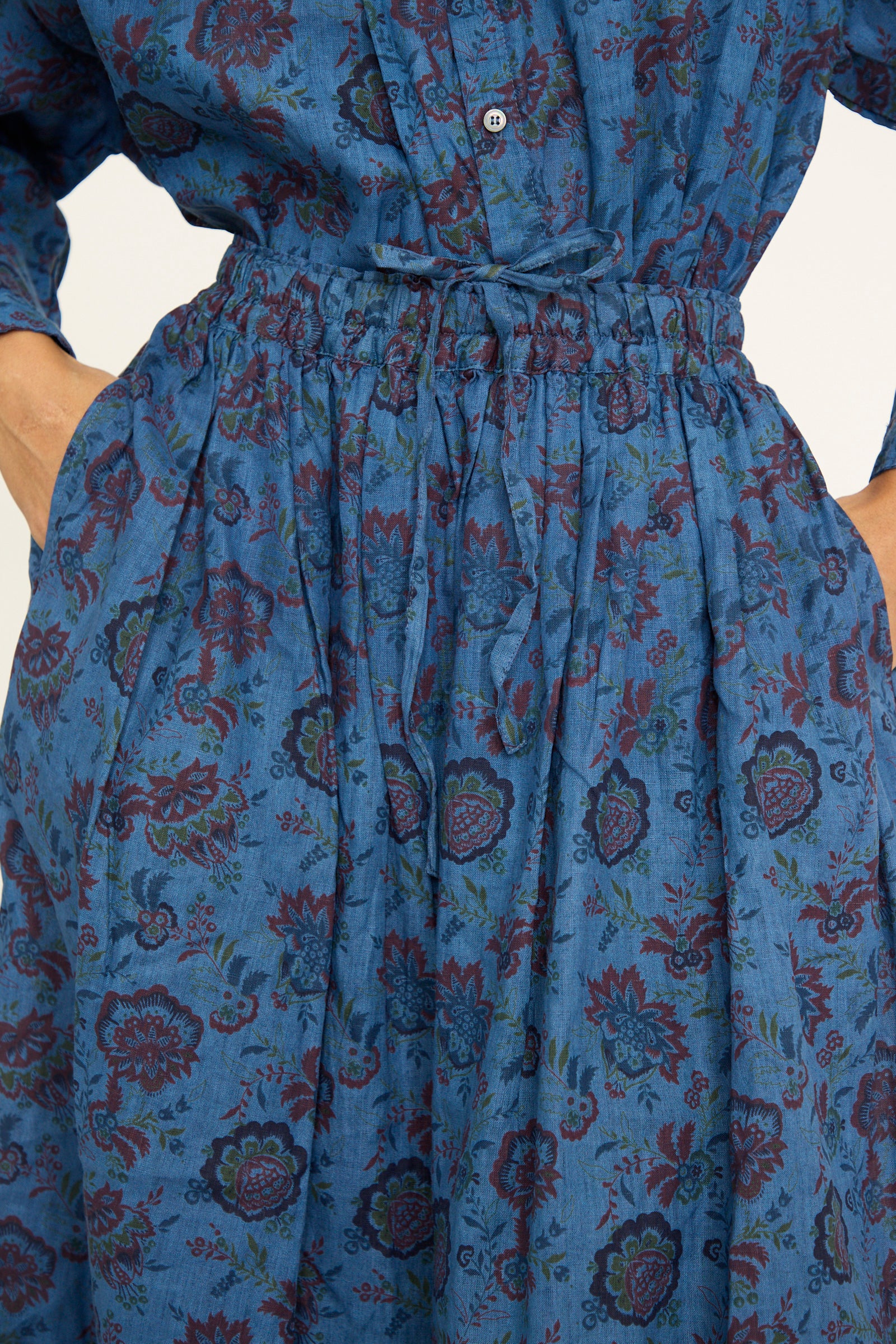 A close-ip of a person wearing an Ichi Antiquités dress in overdye blue with a floral pattern, featuring a long-sleeved design and an A-line silhouette with a high waist and pockets.