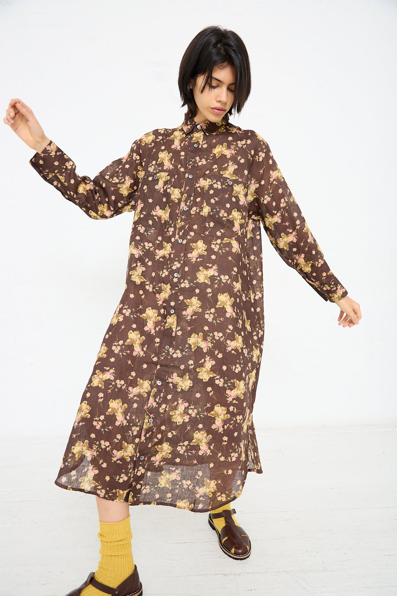 A person stands against a white background wearing the Linen Flower Print Dress in Brown by Ichi Antiquités and yellow socks with brown sandals. They have dark, shoulder-length hair and appear to be adjusting their sleeves.