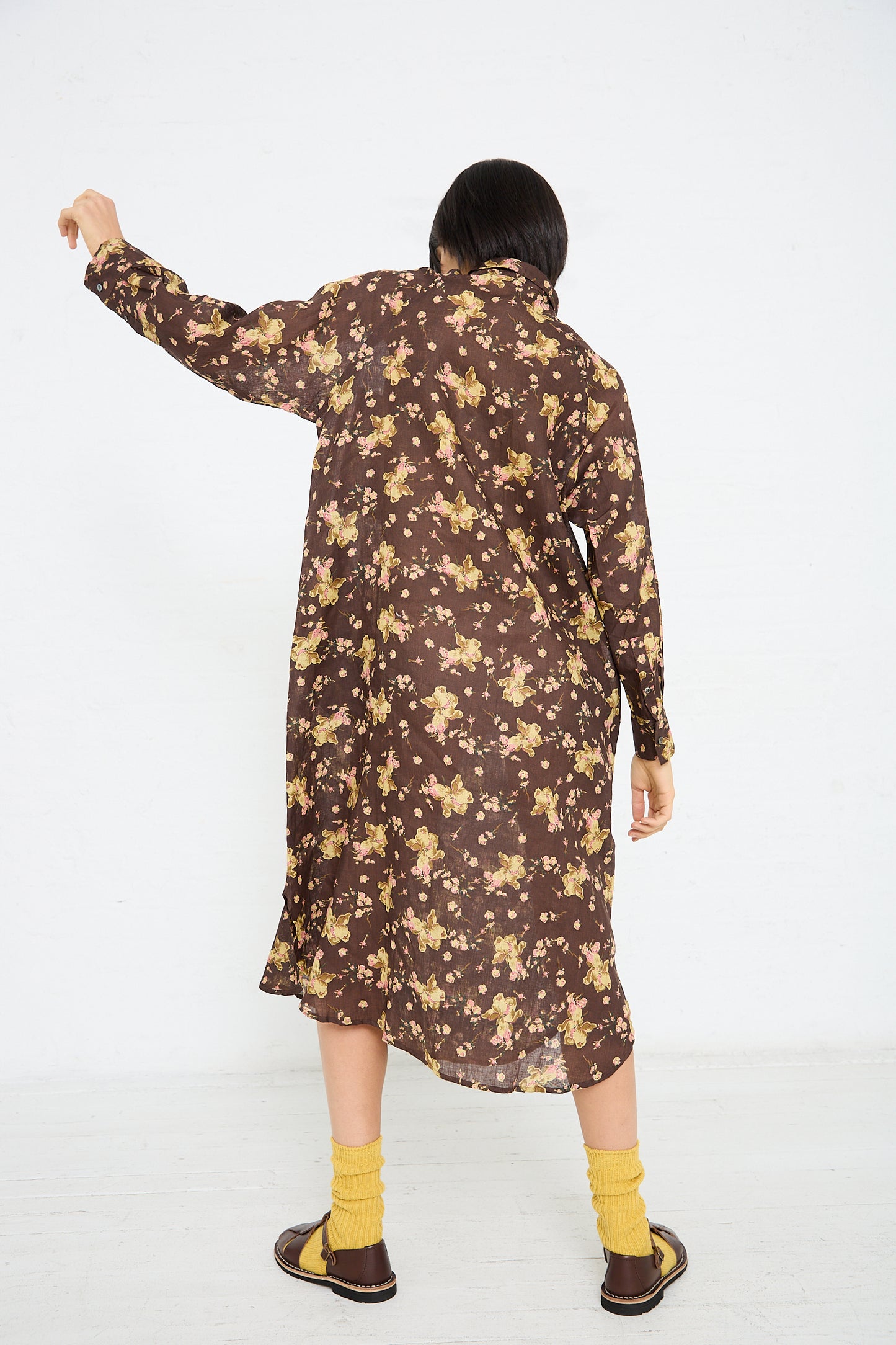 A person stands with their back to the camera, wearing a long, dark brown Linen Flower Print Dress in Brown by Ichi Antiquités, yellow socks, and brown shoes. They are raising one arm.