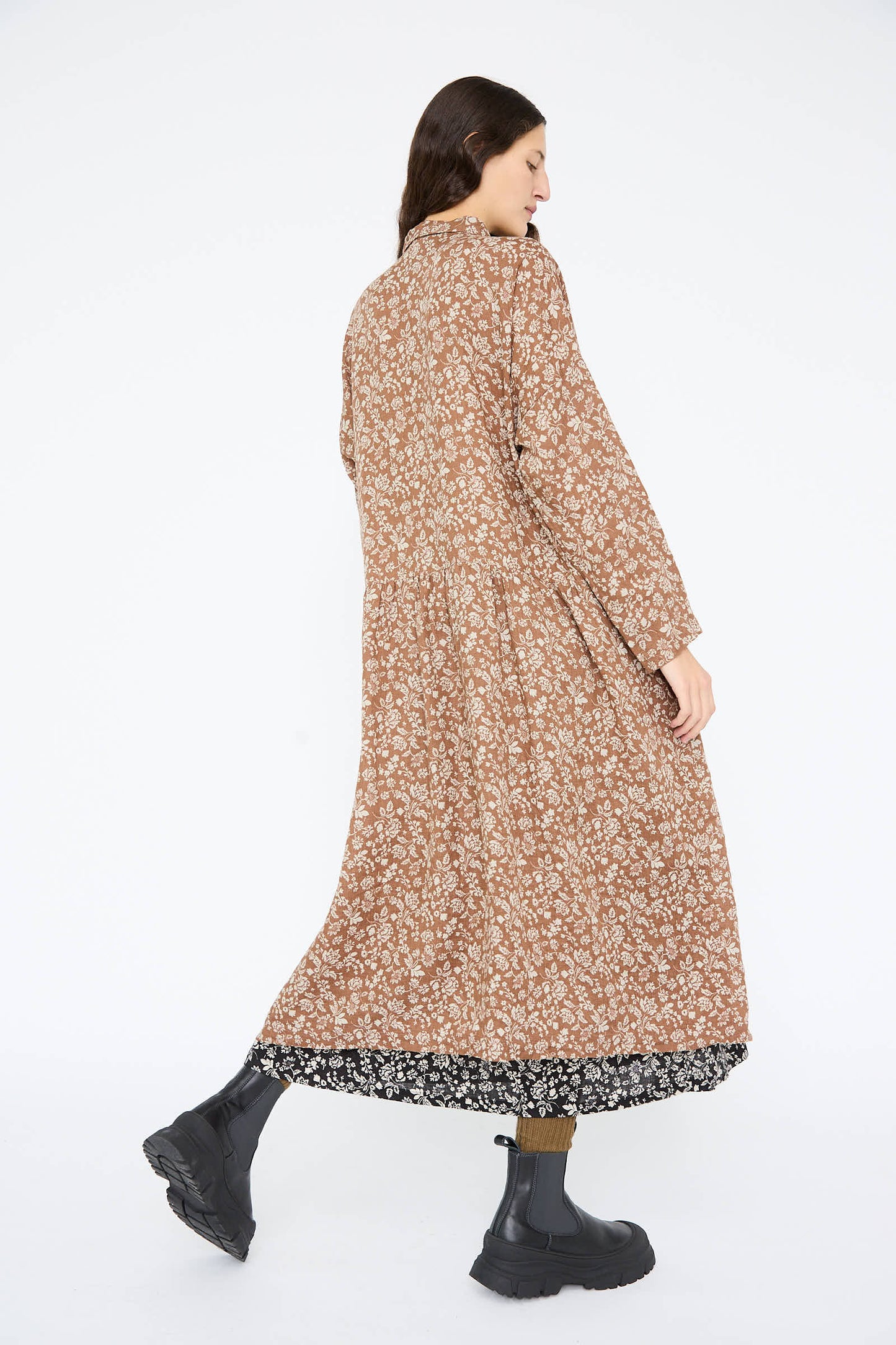 A person models the Ichi Antiquités Linen Flower Print Dress in Brown, featuring a relaxed fit and black floral trim, paired with black boots, while standing turned away on a white background.