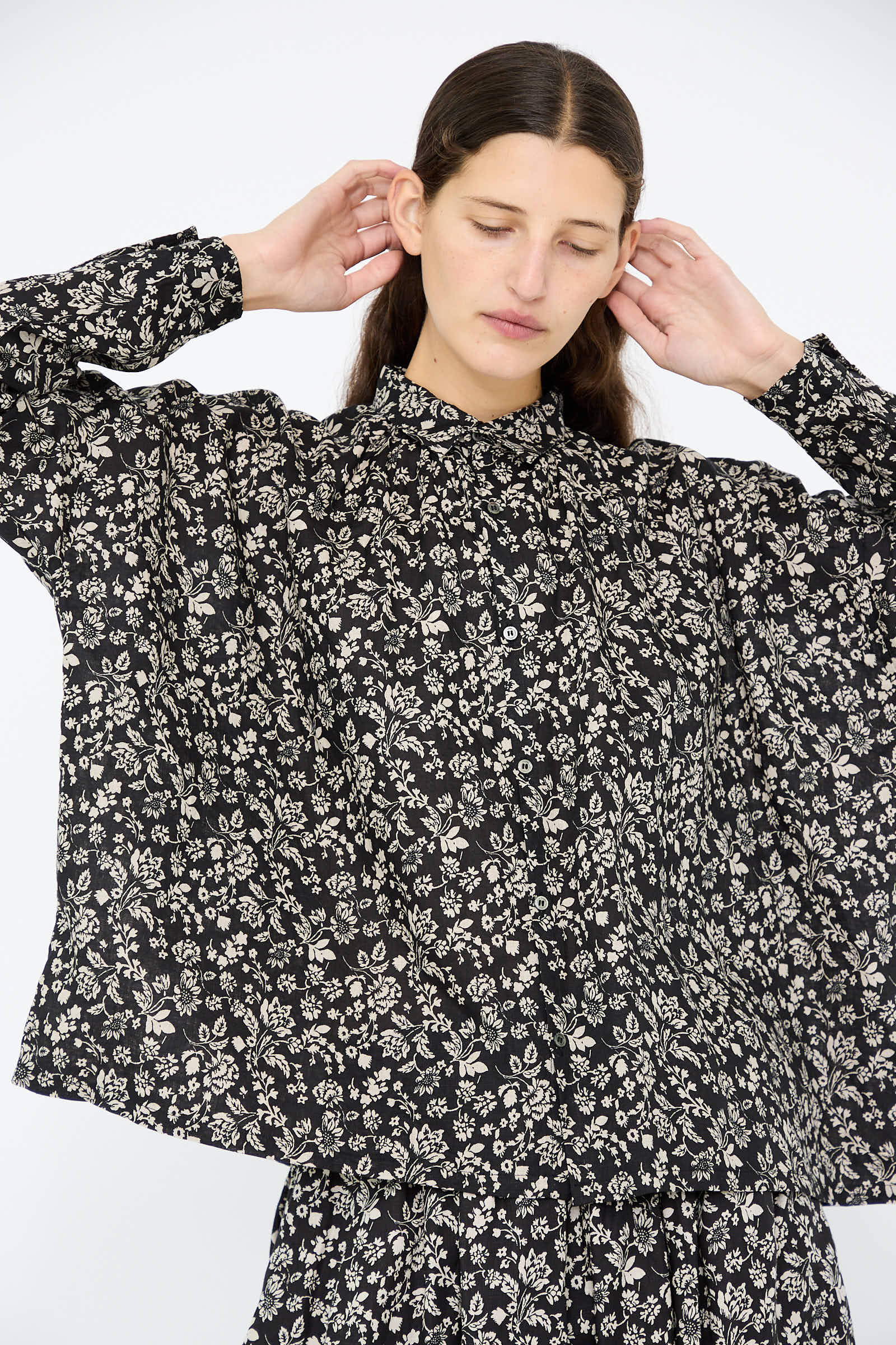 Dressed in a Linen Flower Print Shirt in Black by Ichi Antiquités, a person stands with their eyes closed and hands near their head against a plain background.