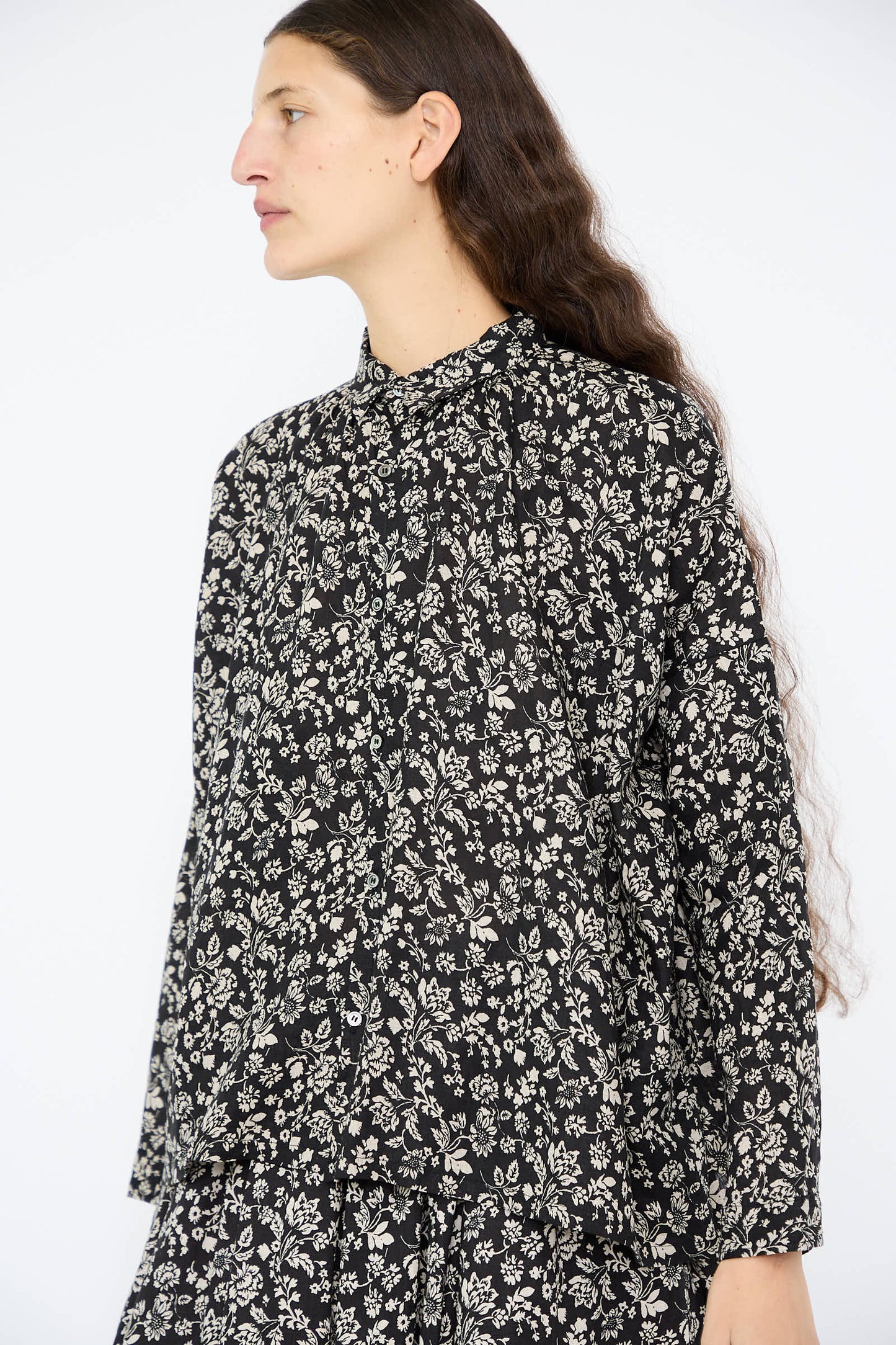 A person wearing the "Linen Flower Print Shirt in Black" by Ichi Antiquités, a one-size, black and white floral-patterned button-up shirt, stands against a plain background.