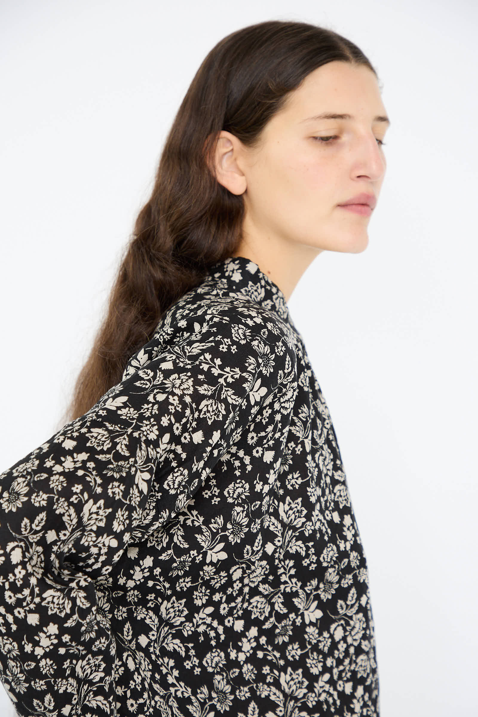 A person with long dark hair wears the Linen Flower Print Shirt in Black by Ichi Antiquités, gazing to the side against a plain background.