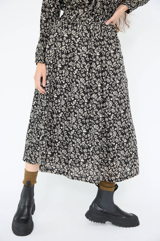 A person is wearing the Linen Flower Print Skirt in Black by Ichi Antiquités, complemented by black boots and brown socks, standing against a plain white background.