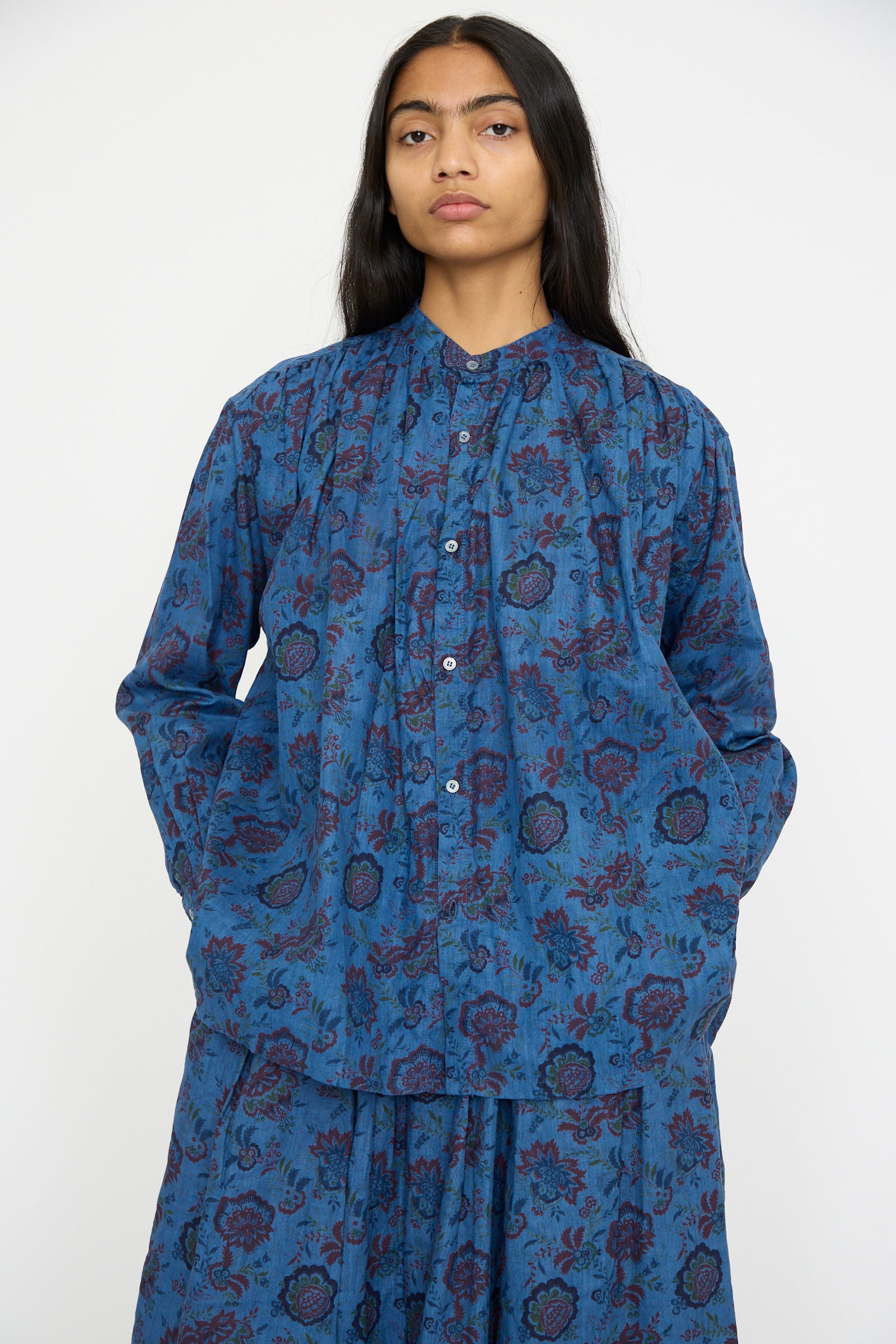 A person models the Ichi Antiquités Linen Flower Shirt in Overdye Blue, a band collar, long-sleeved shirt with a knee-length skirt, against a plain background.