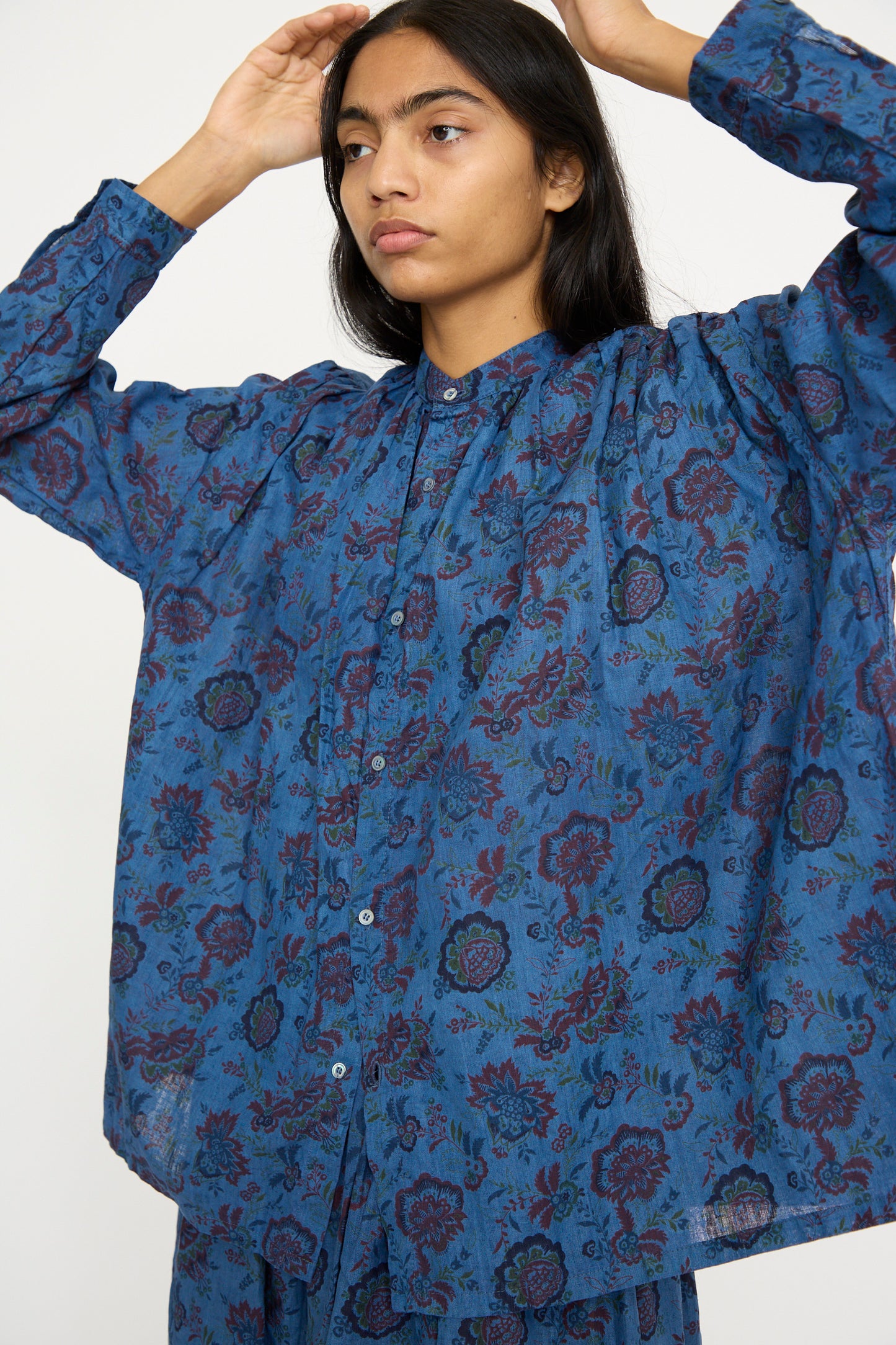 Against a plain background, a person in the Linen Flower Shirt in Overdye Blue by Ichi Antiquités, showcasing a relaxed fit and floral pattern with linen's natural texture, adjusts their hair.