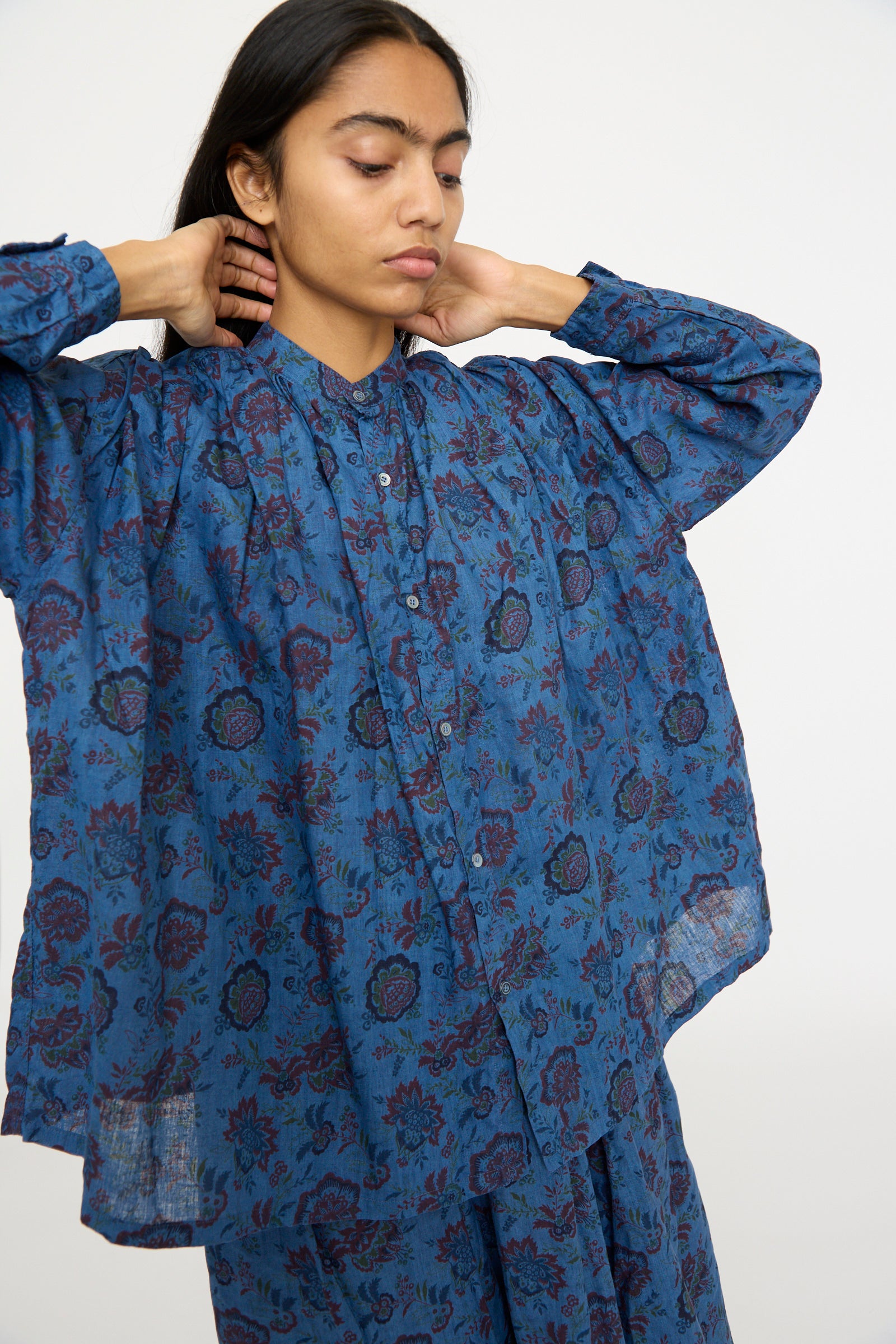 A person wearing the Ichi Antiquités Linen Flower Shirt in Overdye Blue adjusts their collar, showcasing the intricate floral pattern against a plain background.