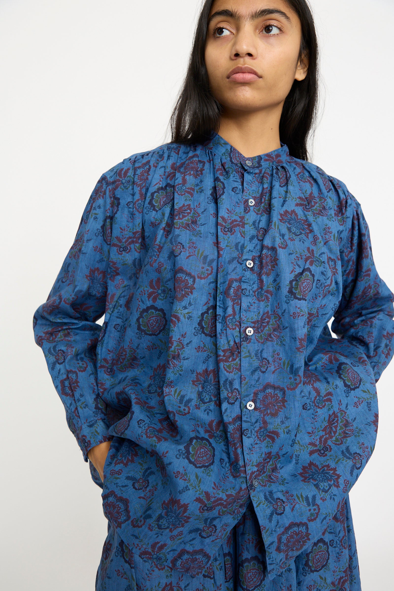 The individual is wearing an Ichi Antiquités Linen Flower Shirt in Overdye Blue, with hands in pockets, glancing away.