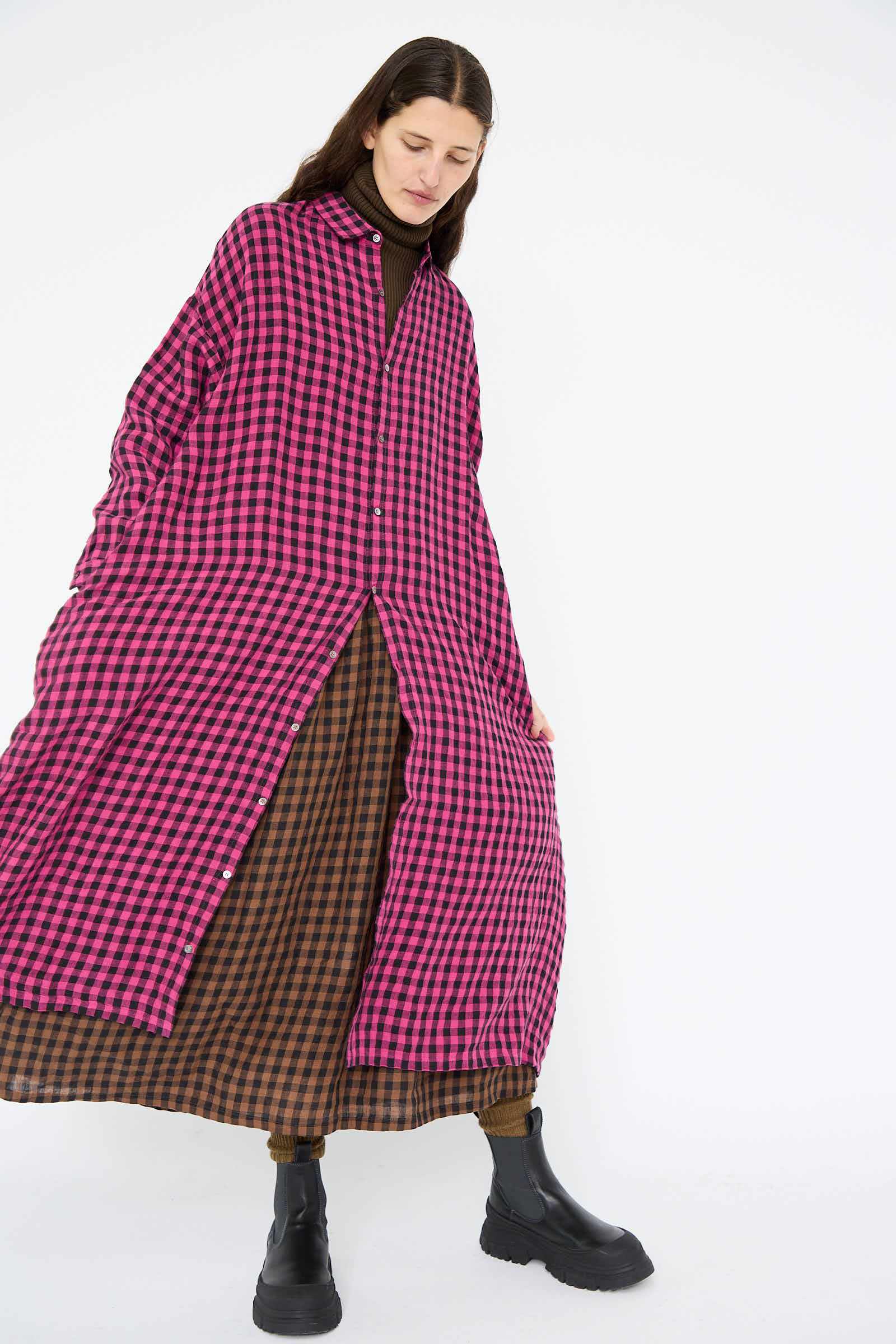 A person wearing the Linen Gingham Check Dress in Pink by Ichi Antiquités over a brown checkered outfit and black boots stands against a plain background.