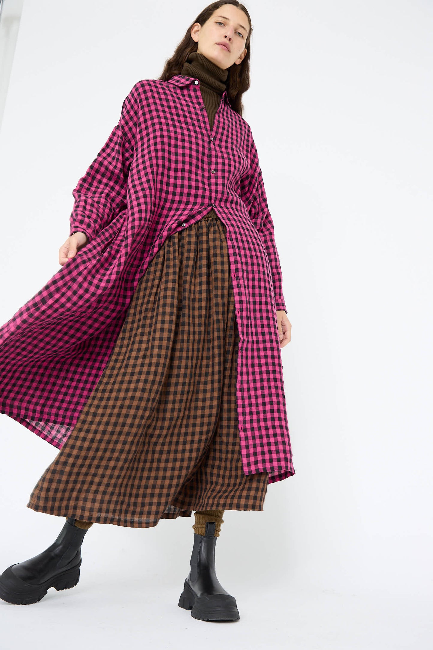 A person wearing the Linen Gingham Check Dress in Pink by Ichi Antiquités, paired with a brown and black plaid skirt, a black turtleneck, and stylish black boots.