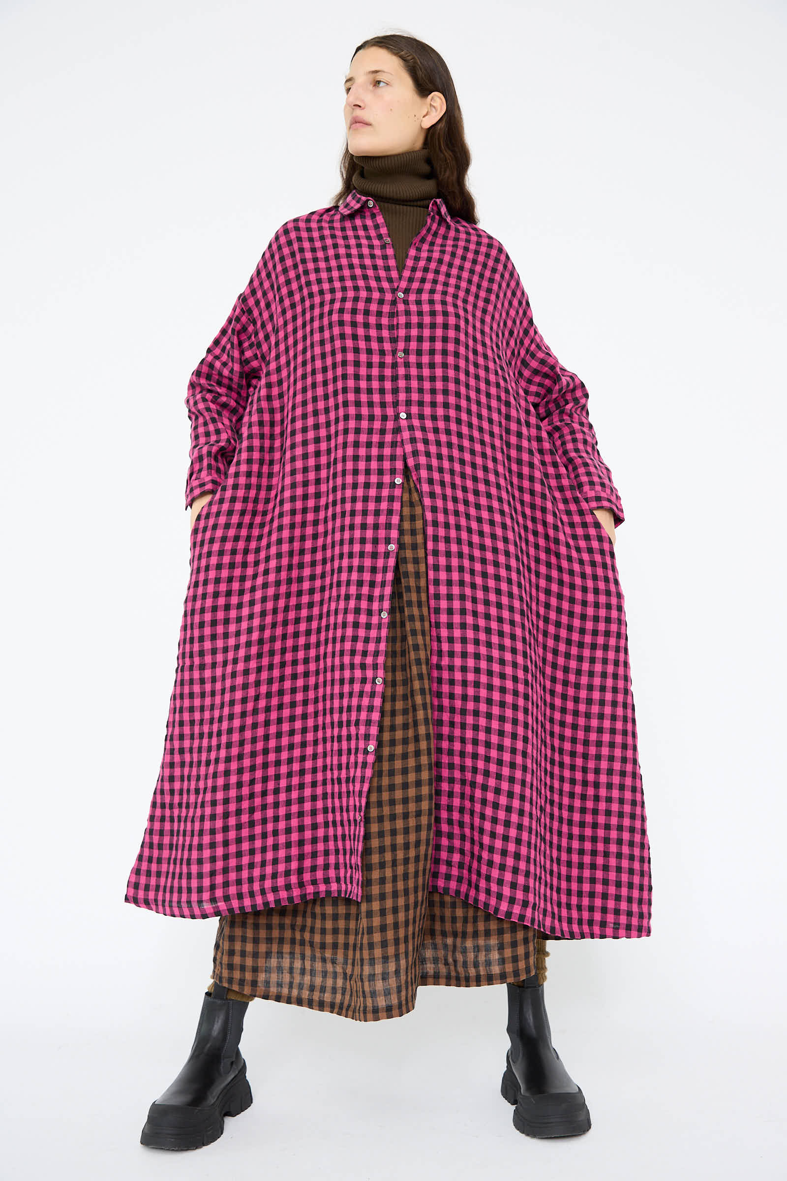 A person dressed in an Ichi Antiquités Linen Gingham Check Dress in pink, paired with brown plaid pants and black boots, is standing against a plain background.