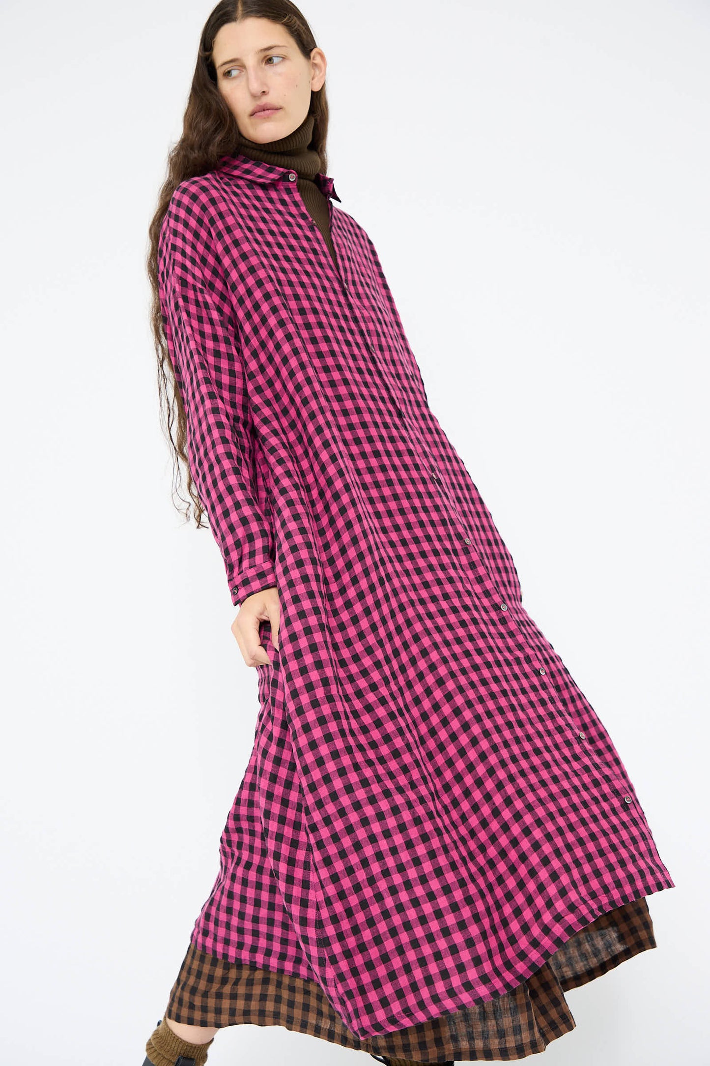 A person wearing the Linen Gingham Check Dress in Pink by Ichi Antiquités, featuring a long design with pink and black checks, layered over a brown turtleneck.