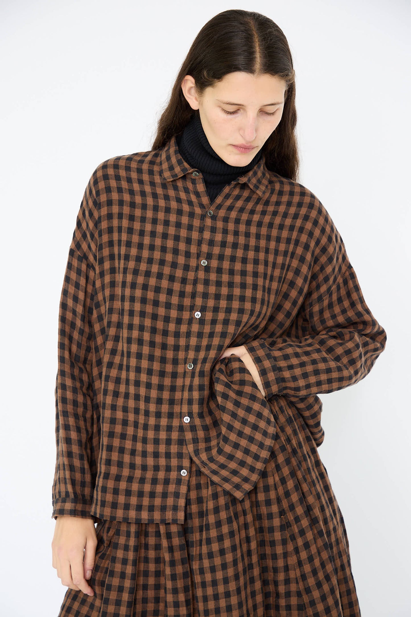 The person wears the oversized Linen Gingham Check Shirt in Brown by Ichi Antiquités over a sleek black turtleneck, gazing downward.