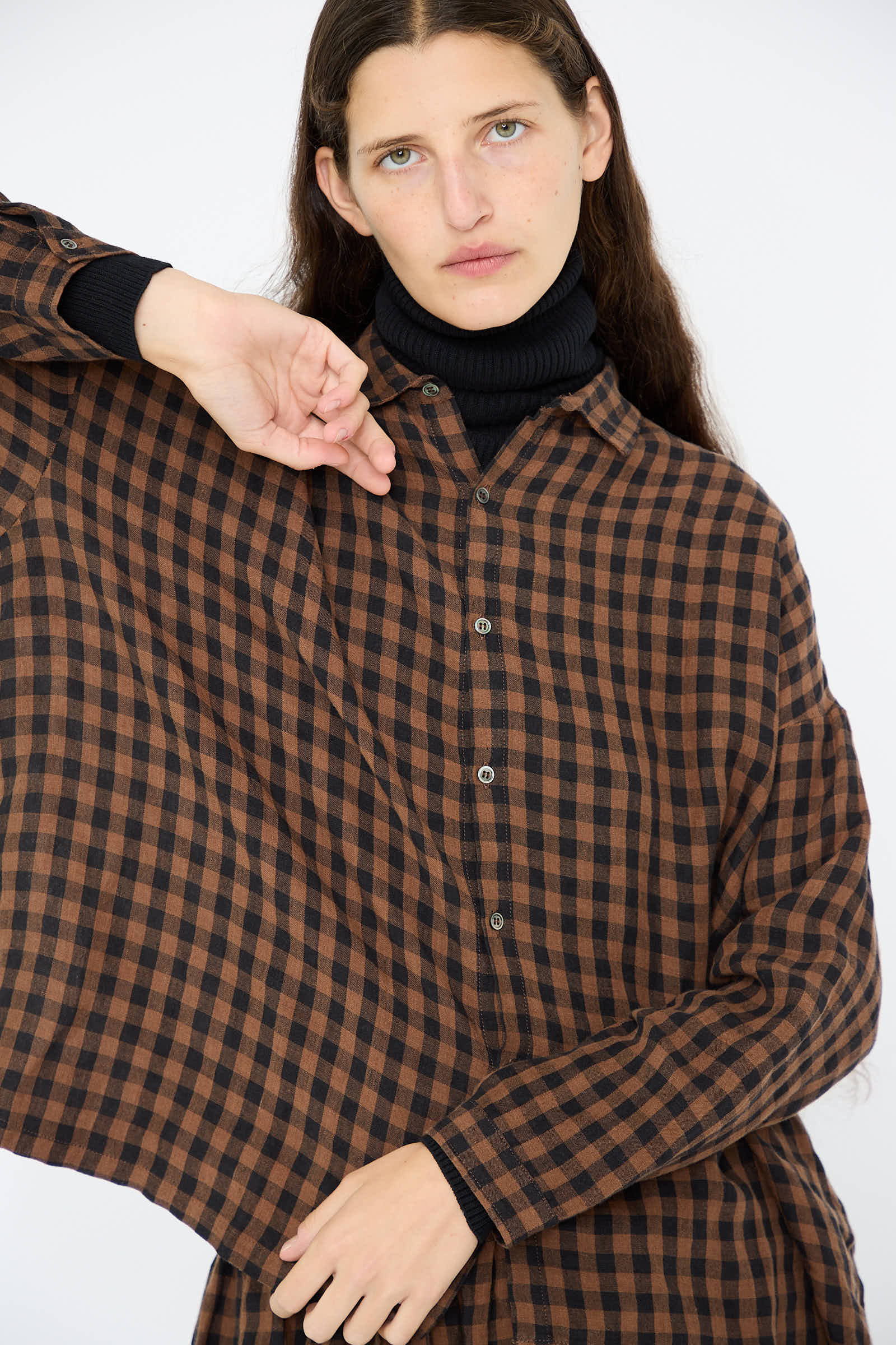 A person wearing the Linen Gingham Check Shirt in Brown by Ichi Antiquités, layered over a black turtleneck, posing with one hand near the collar.