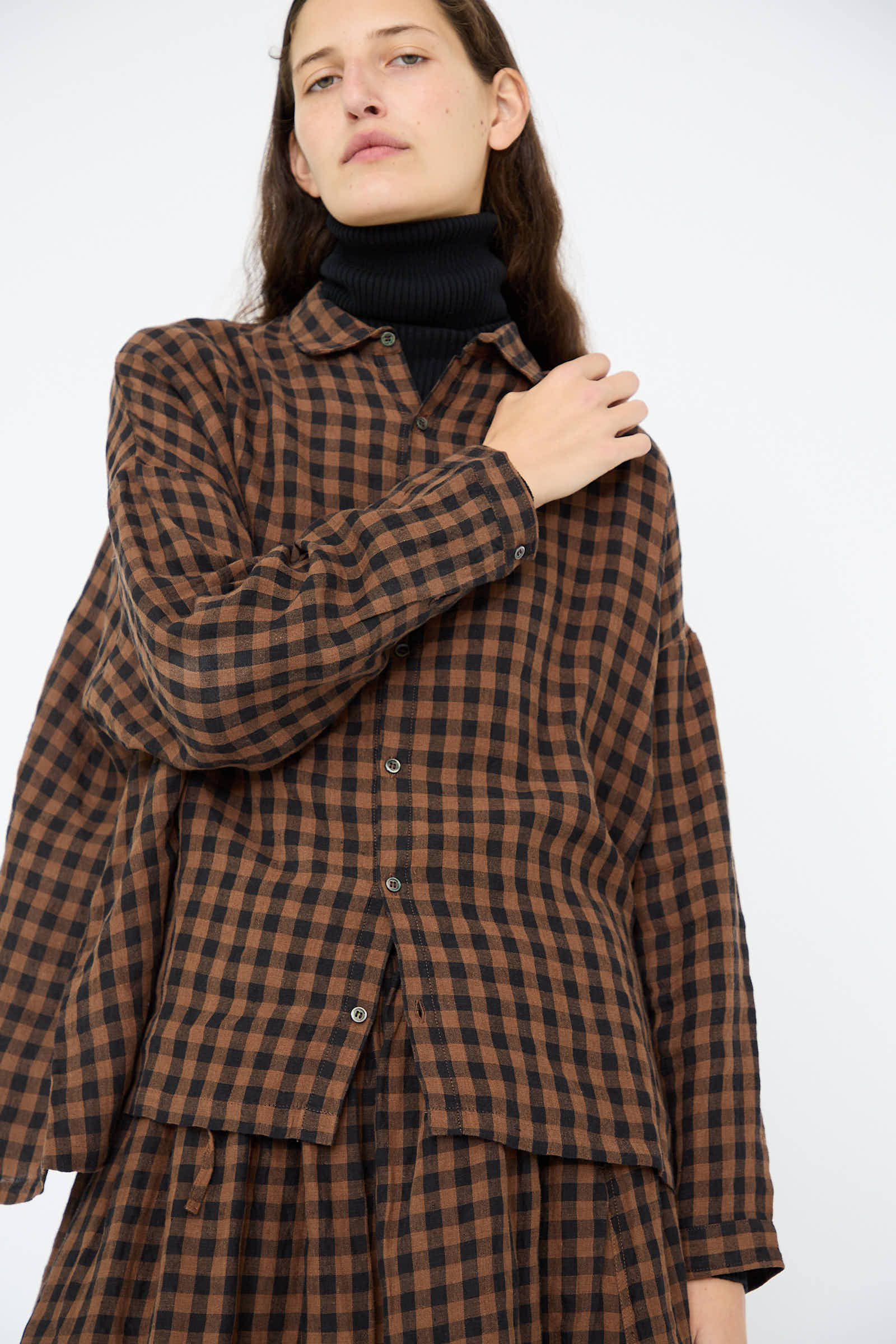 A person in an Ichi Antiquités Linen Gingham Check Shirt in Brown paired with a black turtleneck stands confidently, one hand resting on their shoulder.