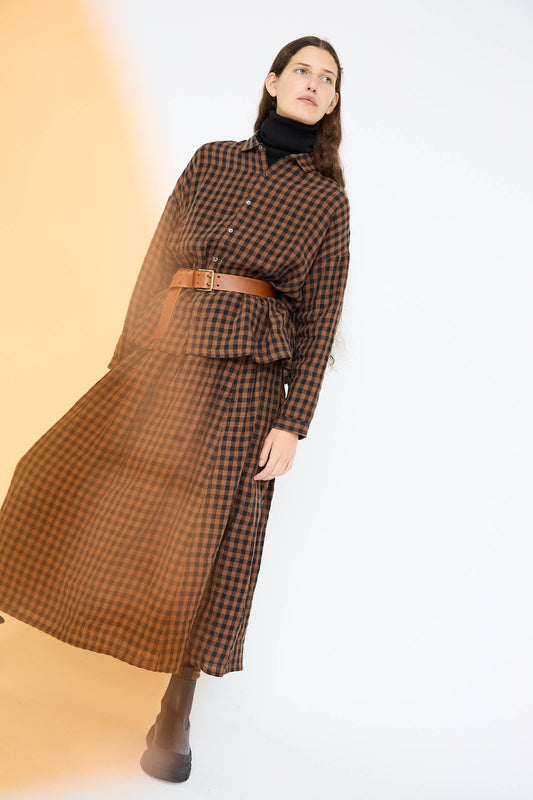 An individual dressed in a brown checkered outfit from Ichi Antiquités, showcasing the Linen Gingham Check Skirt in Brown paired with a black turtleneck, standing against a white background illuminated by a soft orange light effect.