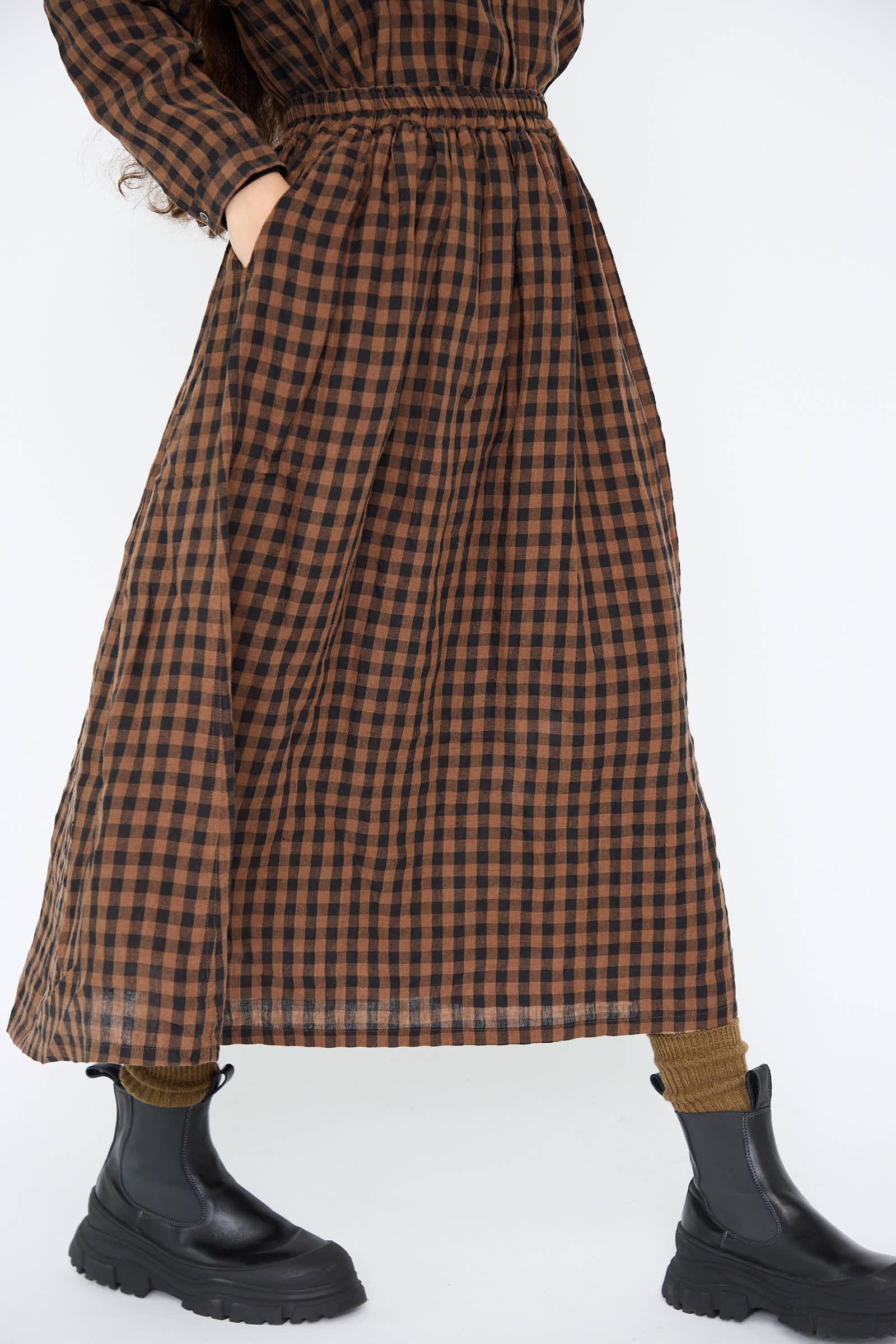 Someone dressed in the Linen Gingham Check Skirt in Brown from Ichi Antiquités, showcasing its elegant A-line silhouette with hands tucked into pockets. The outfit is stylishly completed with black boots and brown socks.