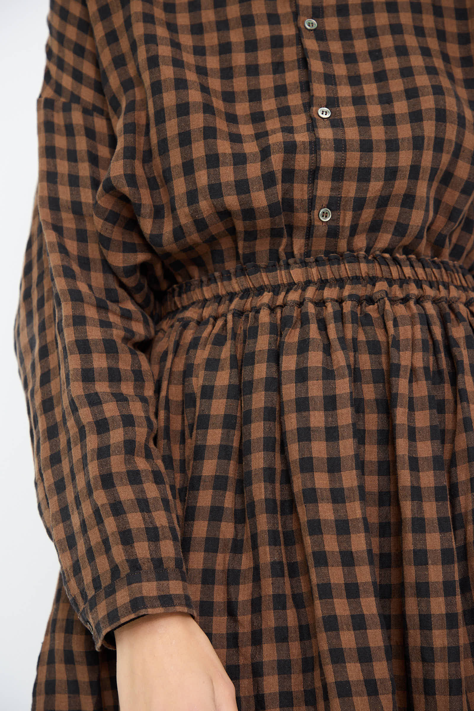 Close-up of a person wearing the Linen Gingham Check Skirt in Brown by Ichi Antiquités. The design showcases a brown and black checkered pattern with buttons down the front and an elastic waist, highlighting its elegant style. Only the torso and hand are visible in the image.