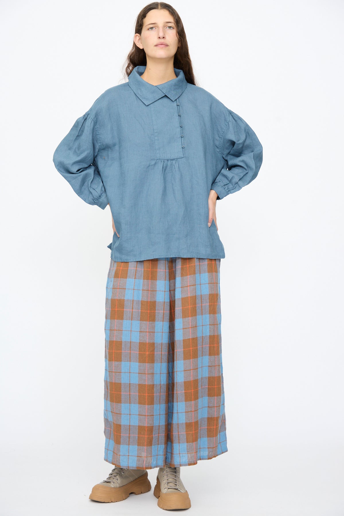 A person wears an Ichi Antiquités Linen Shirt in Earth Blue with puffed sleeves, wide-leg blue and brown plaid trousers, and tan platform sneakers.