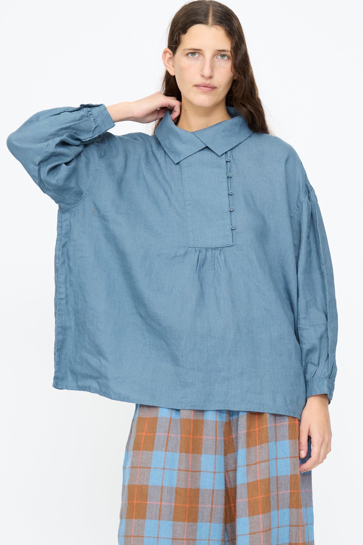 A person wearing an Ichi Antiquités Linen Shirt in Earth Blue, a relaxed-fit cotton linen top with a pleated bodice, paired with a blue and orange plaid skirt.
