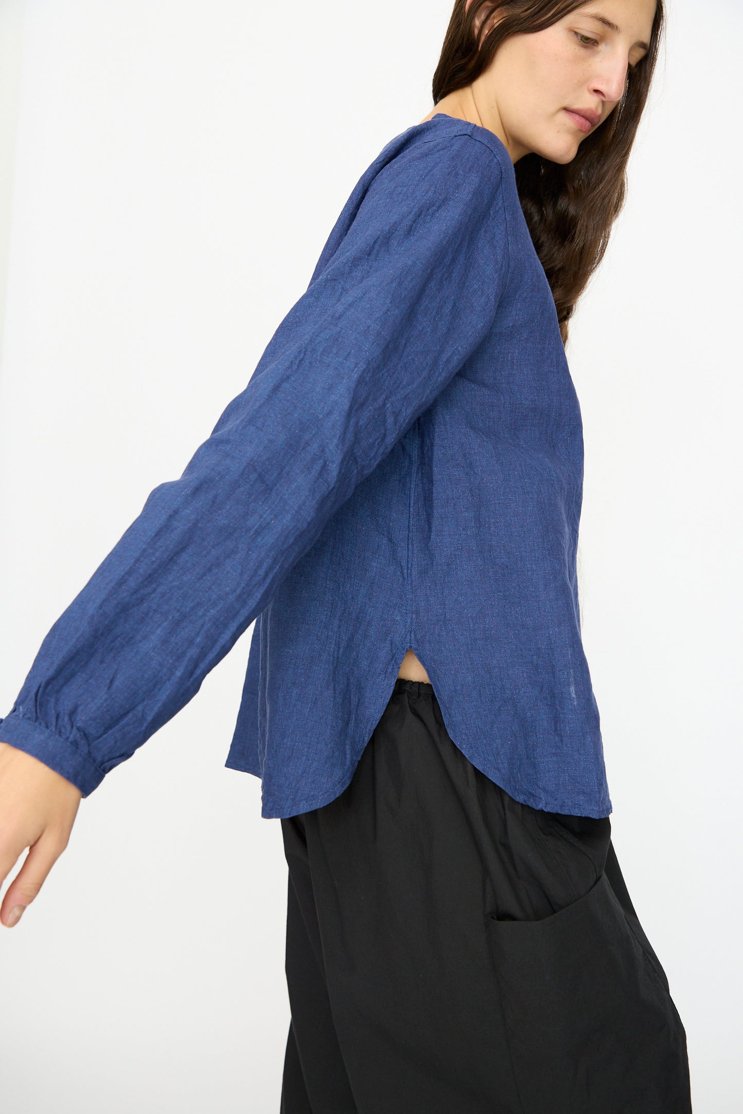 A person models the Ichi Antiquités Linen Shirt in Indigo with loose black pants, standing sideways against a plain white background and extending one arm.