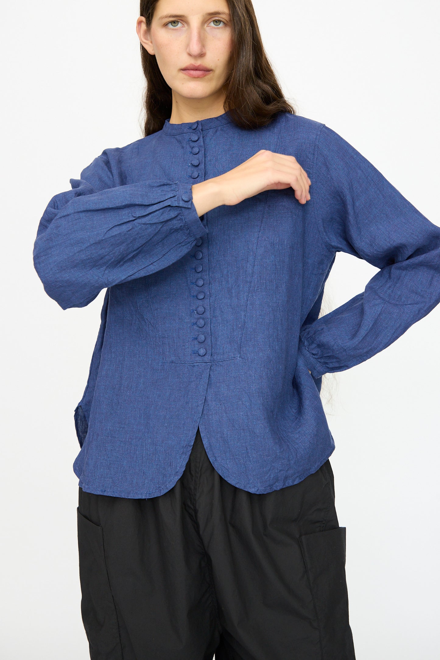 A person models the Ichi Antiquités Linen Shirt in Indigo, featuring a band collar, paired with black pants, standing against a plain background with one arm bent and the other hand on their hip.